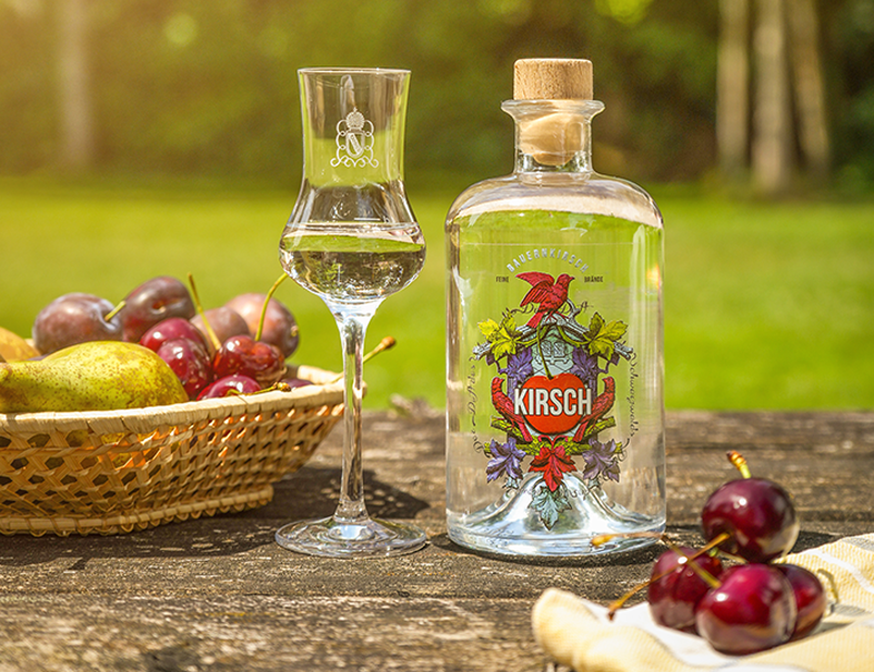 Bauernkirsch-Flasche mit Obstbrand und gefülltem Glas neben einem Obstkorb und frischen Kirschen auf einem Holztisch im Garten.
