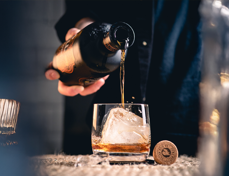 Hand schenkt einen bernsteinfarbenen Likör oder Whisky aus einer dunklen Flasche in ein Glas mit Eiswürfel.