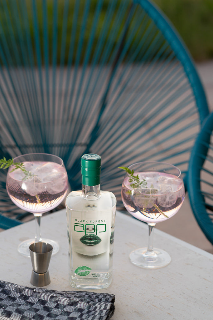 Eine Flasche Black Forest Gin neben zwei Gin Tonic Gläsern mit Kräutergarnitur, auf einem Tisch im Freien.