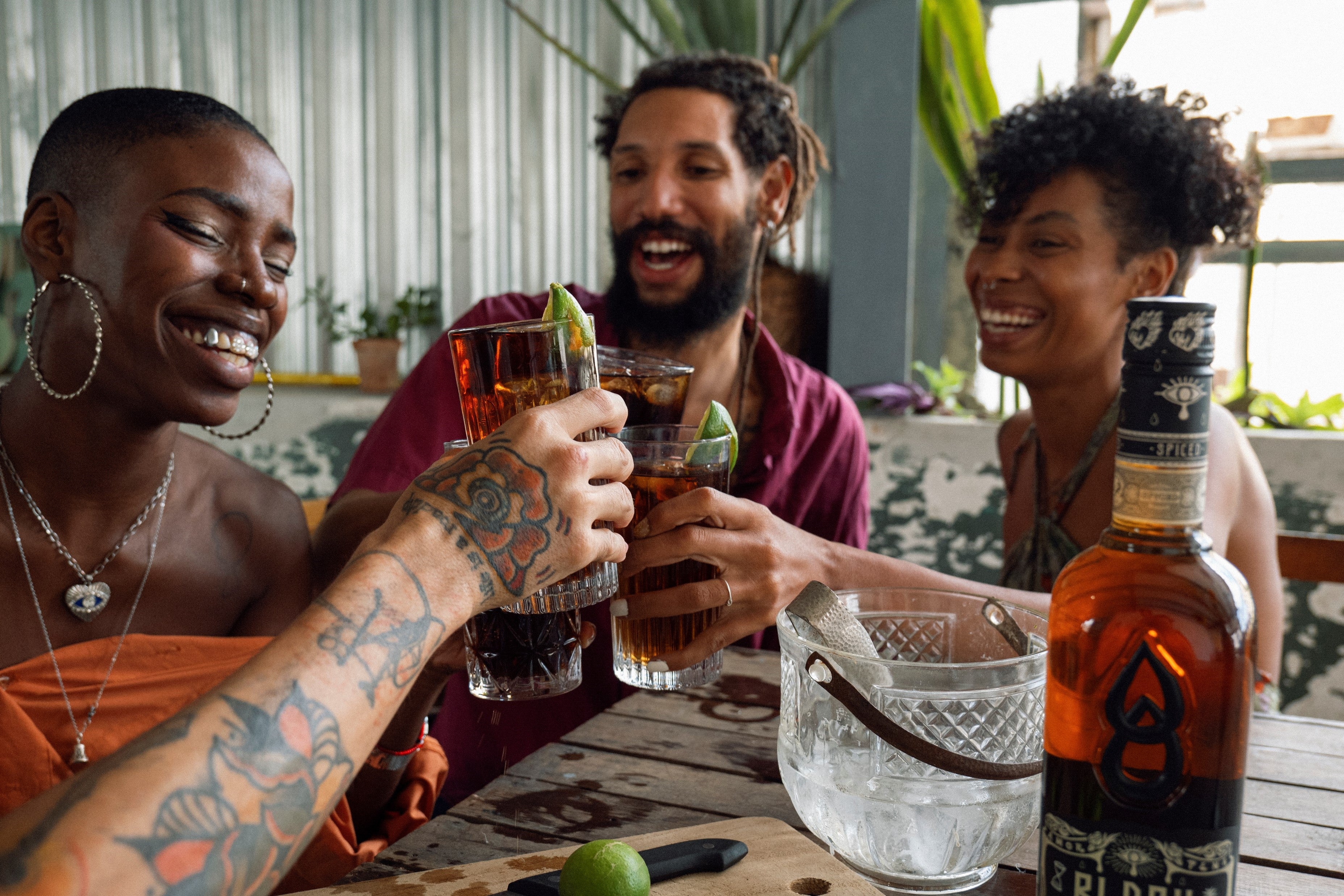 Drei lachende Menschen stoßen mit Longdrinks an einem Tisch mit Benutz, Flasche und Messer darauf an.