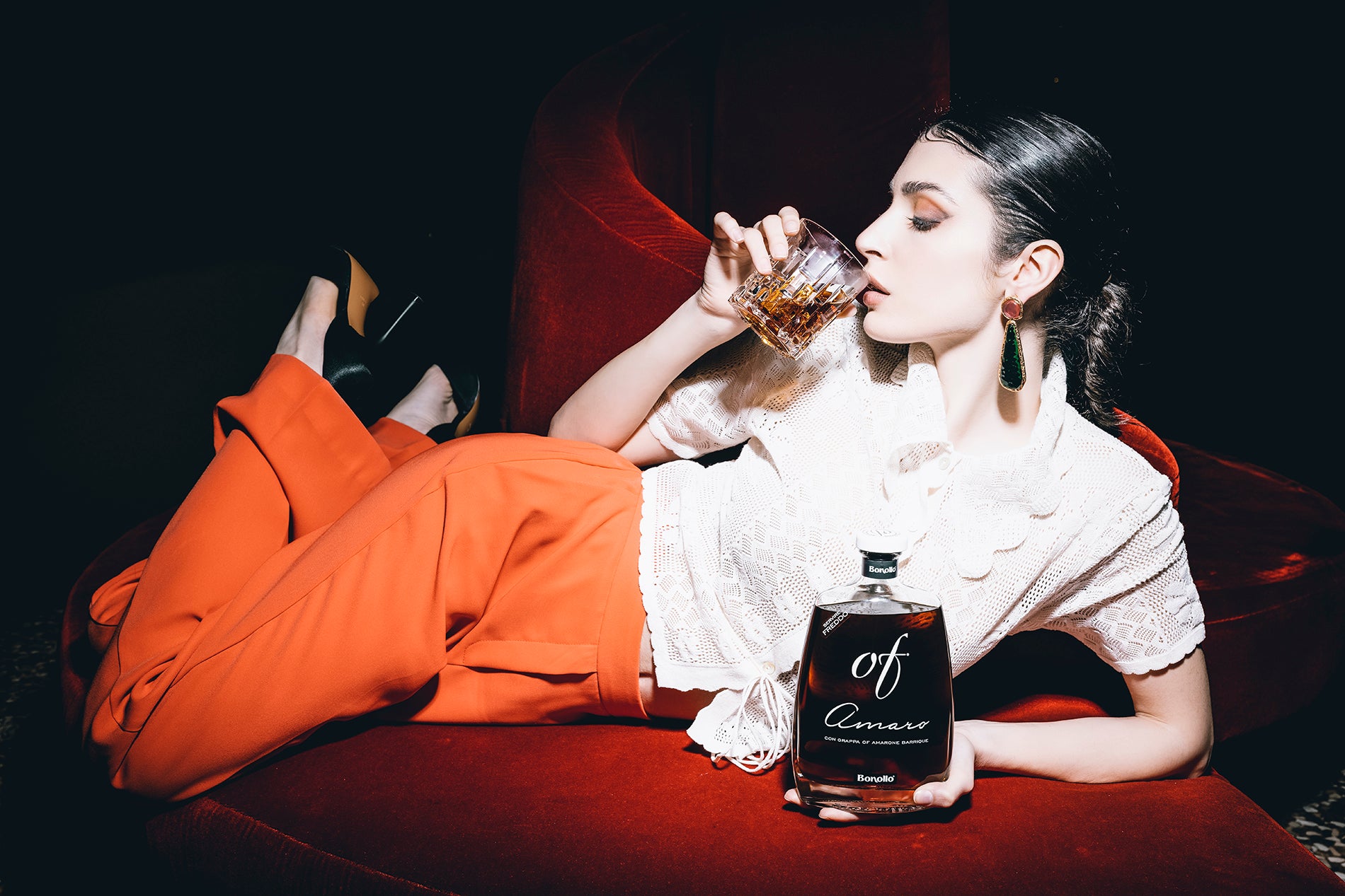 Frau in weißem Spitzenoberteil und orangefarbener Hose, trinkend aus einem Glas auf rotem Sofa mit Flasche Amaro.