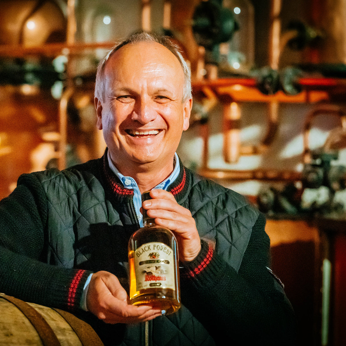 Ein Mann hält eine Flasche "Black Forest" Schnaps lächelnd in einer Brennerei.