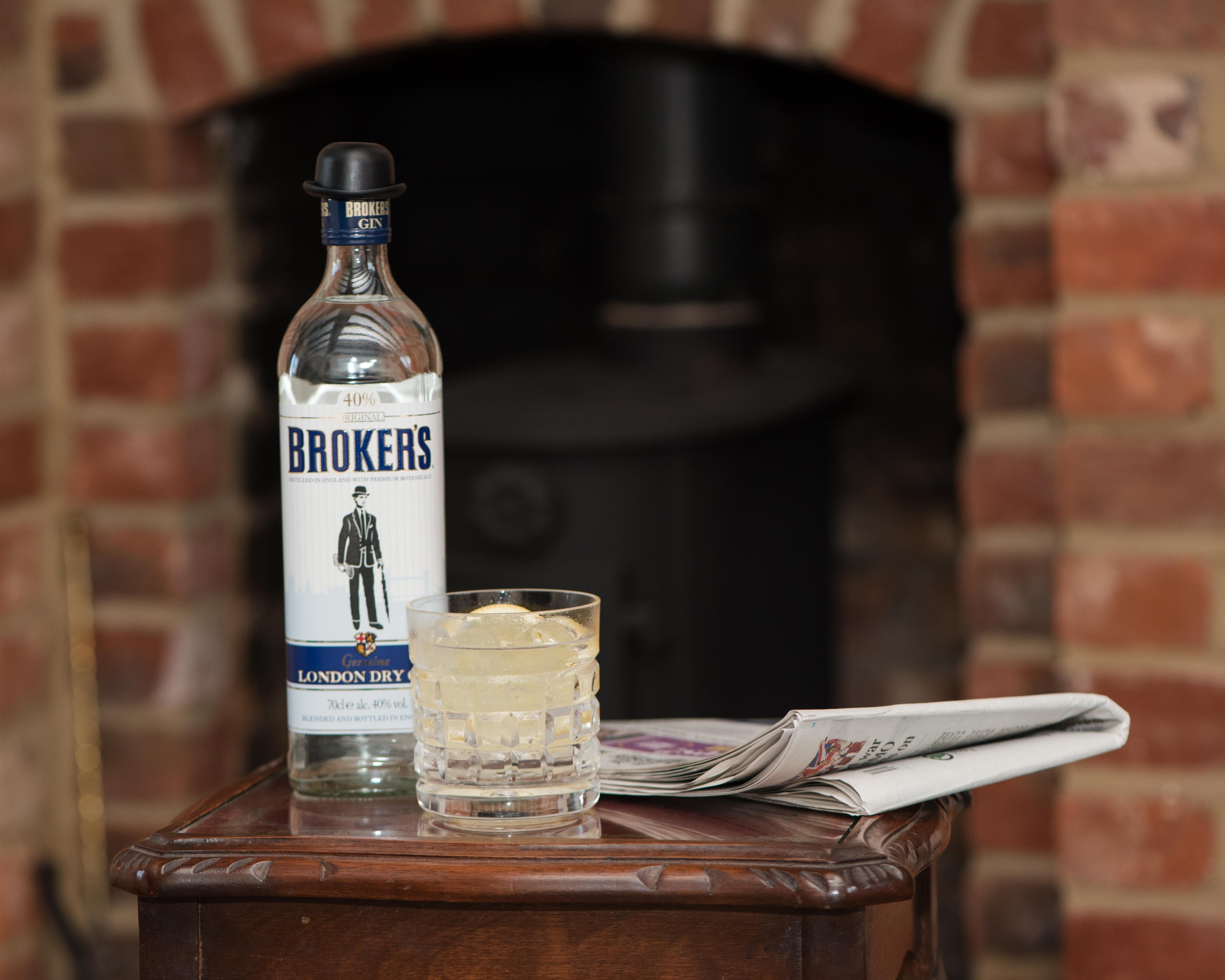 Eine Flasche Gin, ein Glas mit Eis und eine Zeitung stehen auf einem Holztisch vor einem gemauerten Kamin.