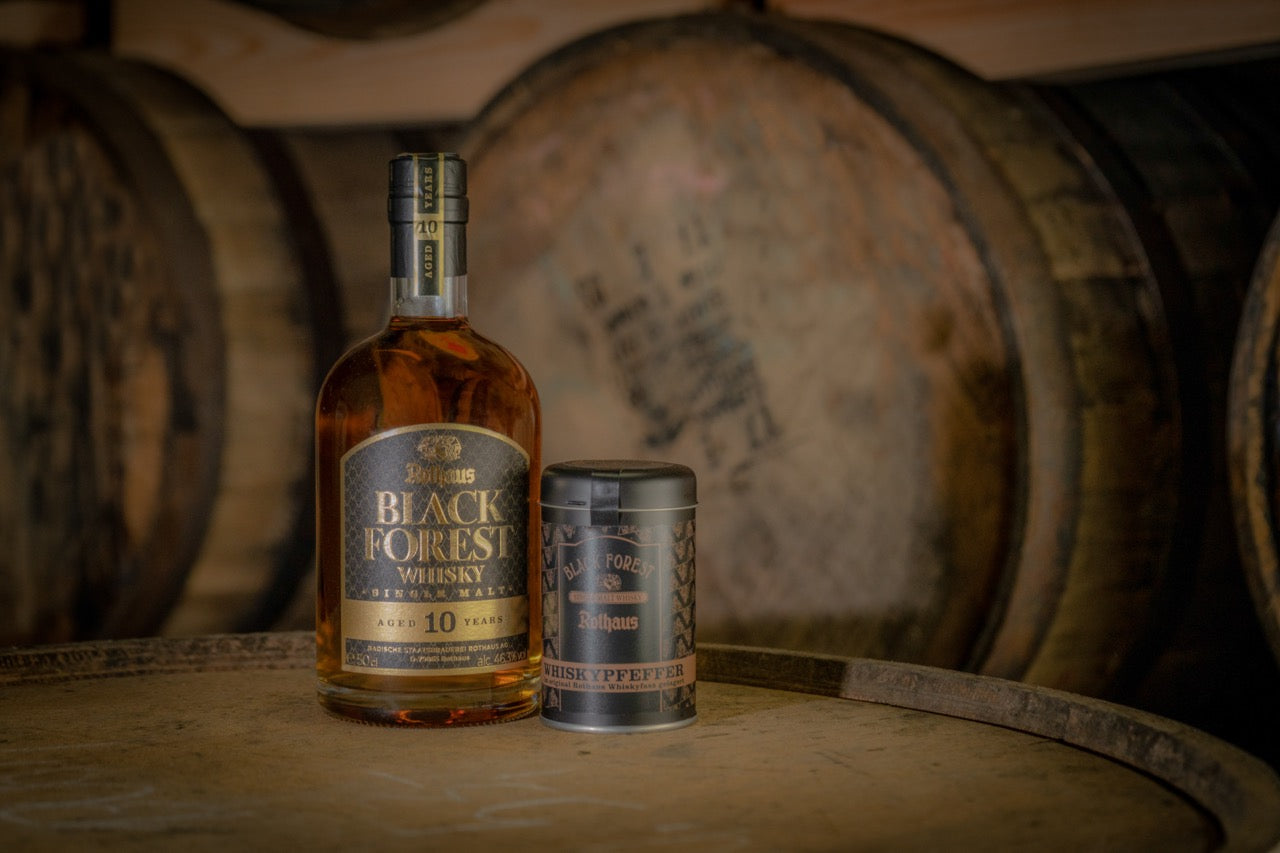 Eine Flasche Black Forest Whisky und eine Dose Whisky-Pfeffer stehen vor Holzfässern.