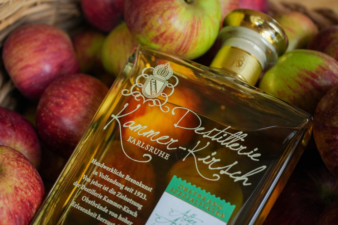 Nahaufnahme einer Flasche Kammer-Kirsch Obstbrand in einem Korb mit roten und grünen Äpfeln.