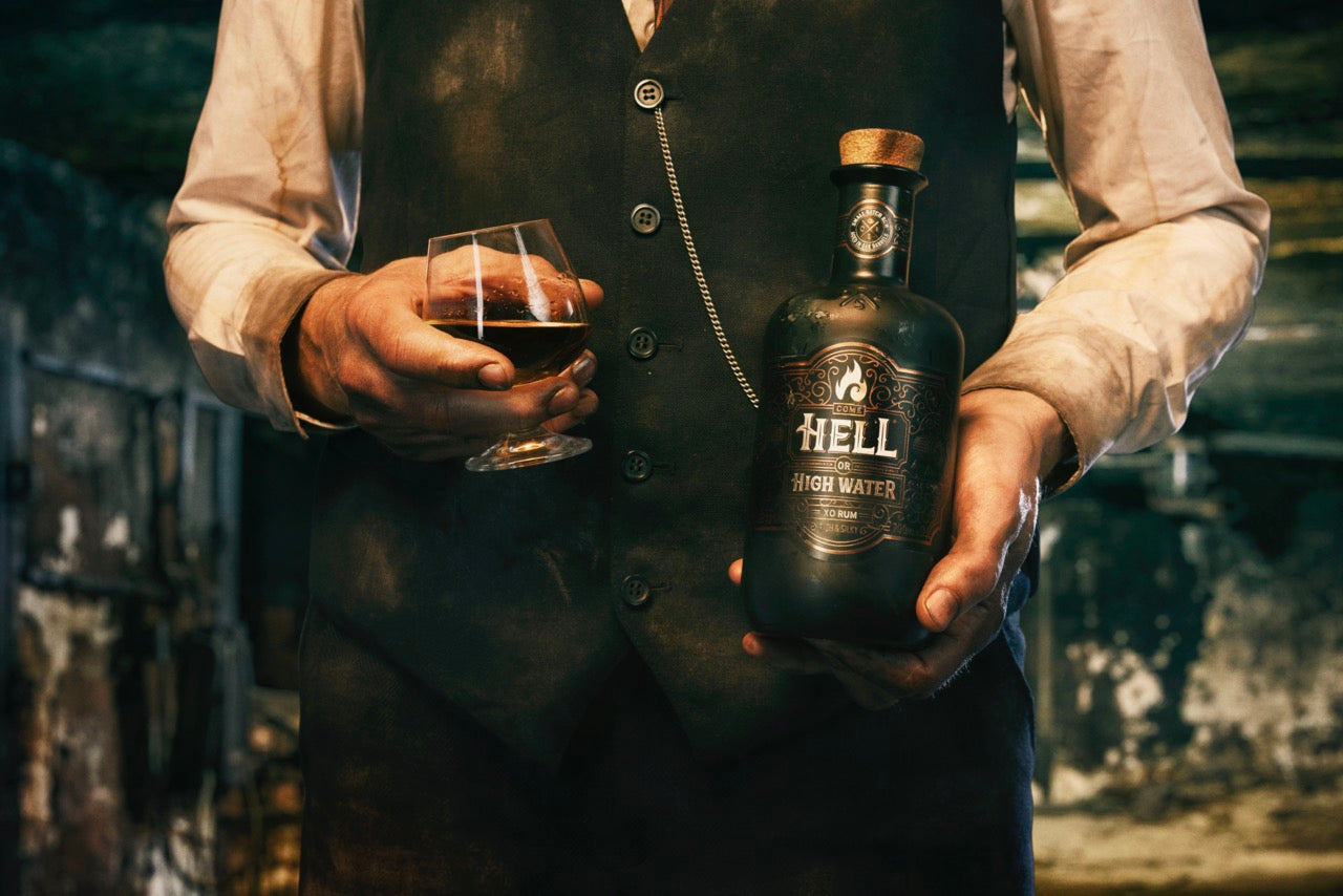 Person hält schwarze Rum-Flasche mit "Hell or High Water" und ein Glas in einer rustikalen Umgebung.