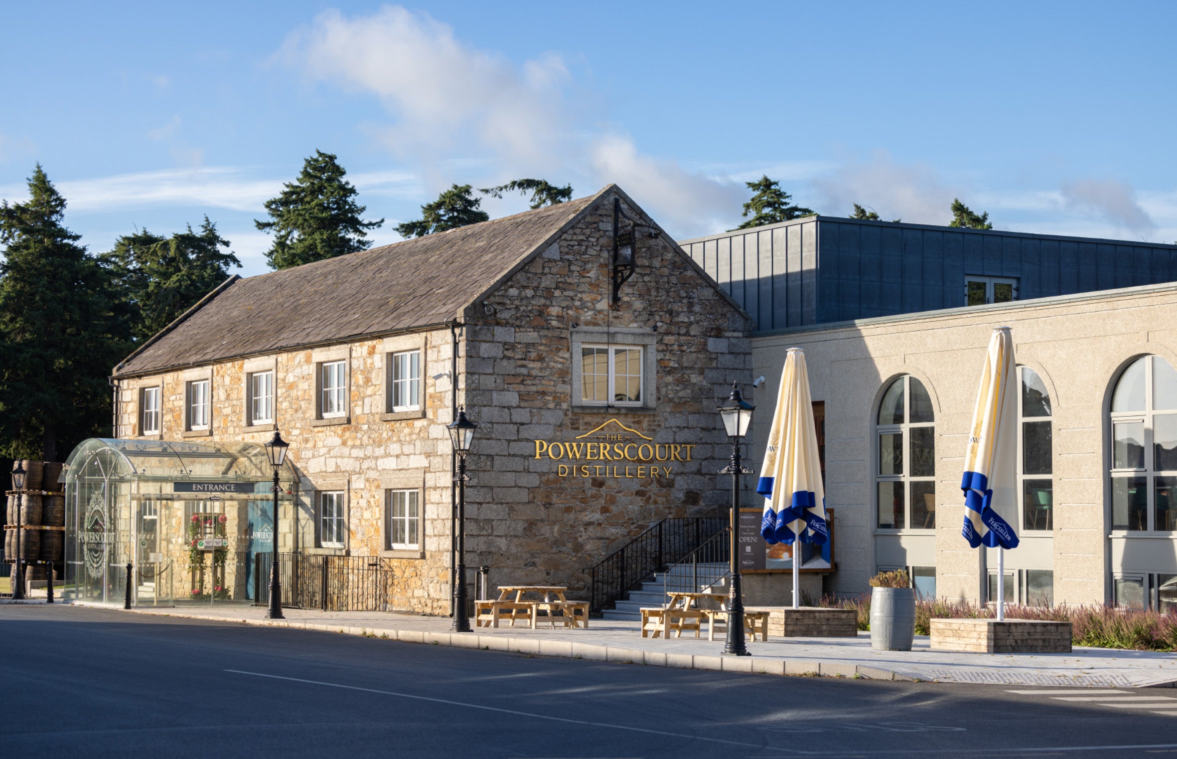 Stein- und Glasgebäude der Powerscourt Distillery mit Schildern und Sonnenschirmen im Freien.
