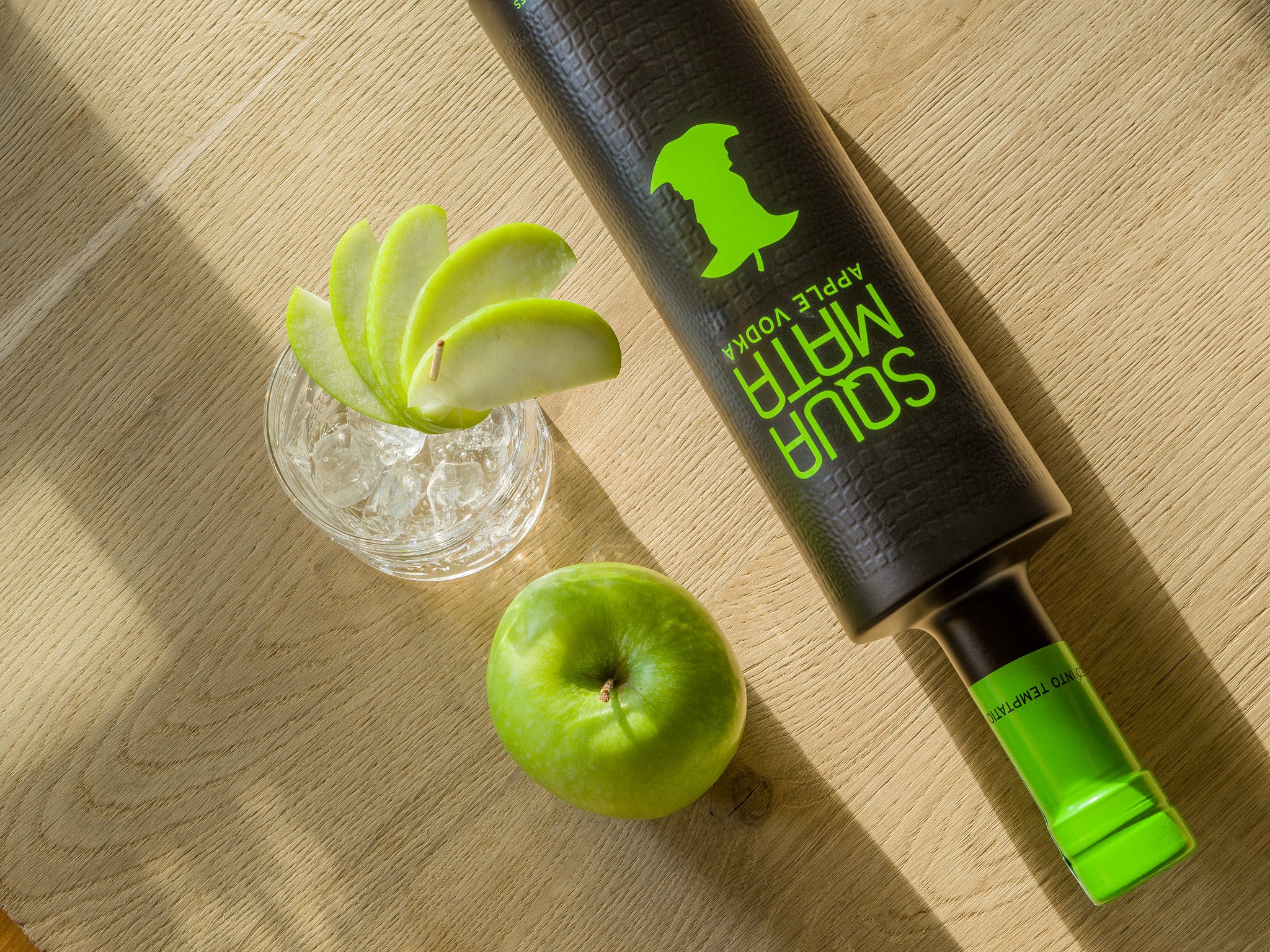 Eine Flasche Apfel-Wodka, ein Glas mit Eis und Apfelscheiben, und ein grüner Apfel auf einem Holztisch.