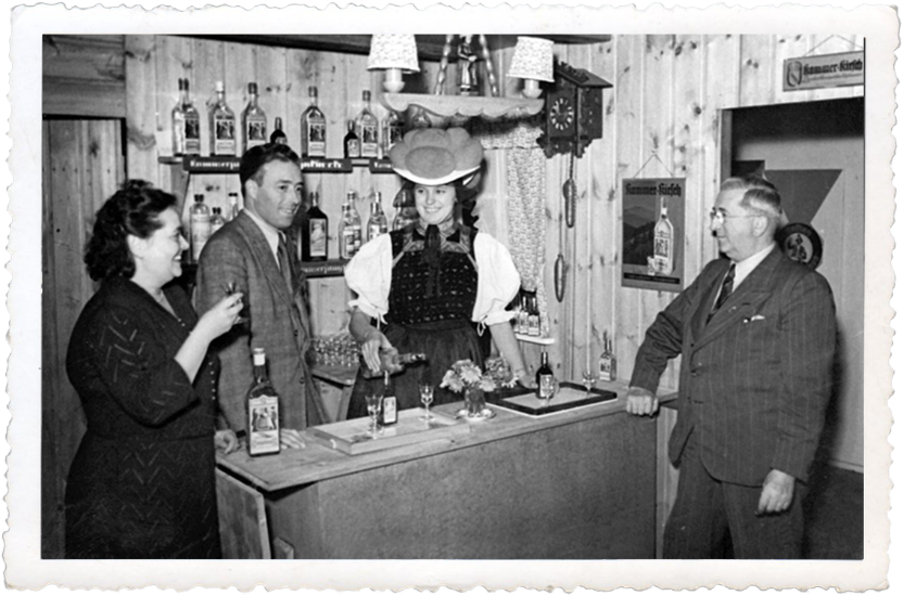 Vier Personen an einer rustikalen Bar mit Alkoholflaschen, eine Frau in Tracht schenkt Getränke ein.