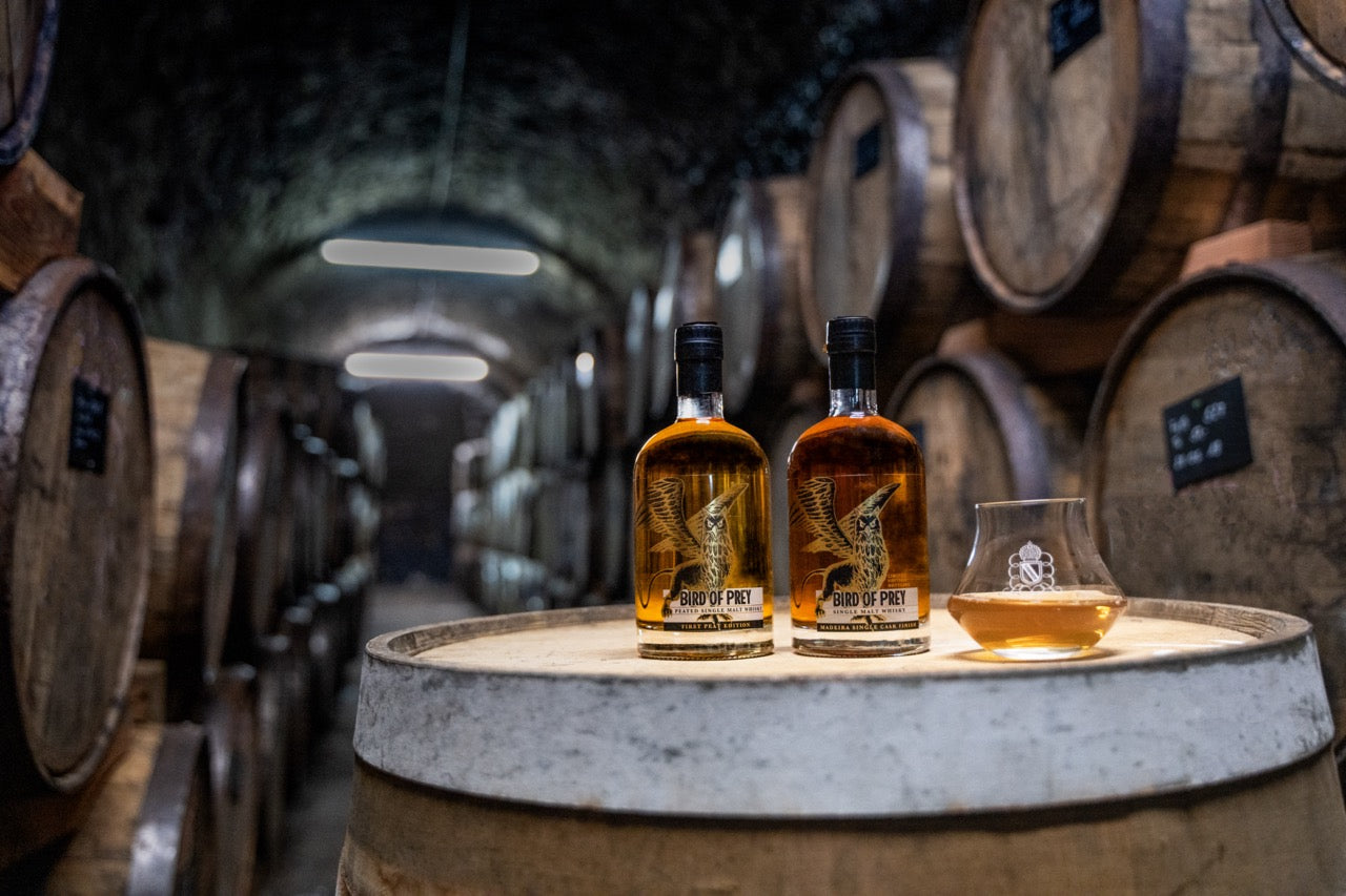 Zwei Whiskyflaschen und ein Glas auf einem Fass in einem Lagerraum mit Holzfässern.