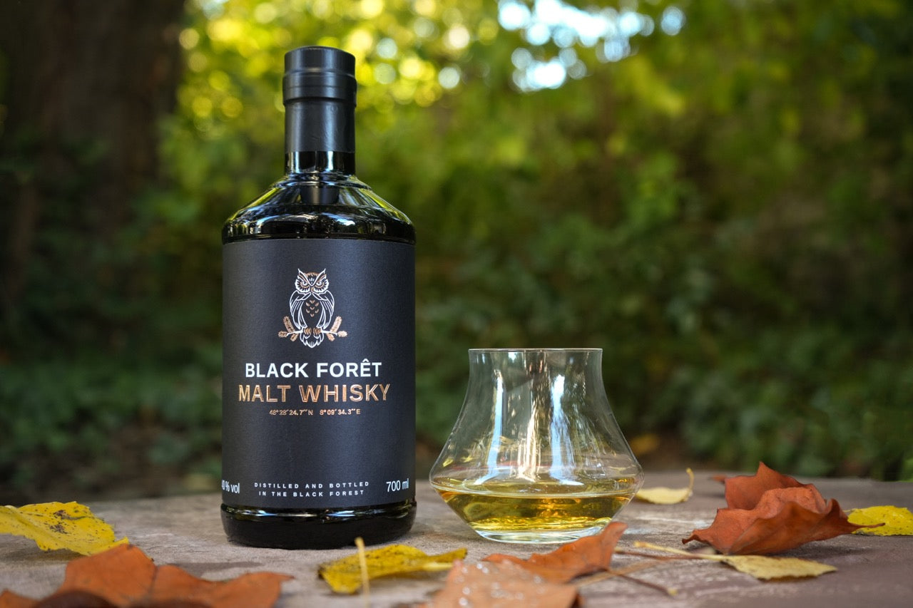 Flasche Black Forêt Malt Whisky und ein Glas mit Whisky, umgeben von bunten Herbstblättern im Freien.
