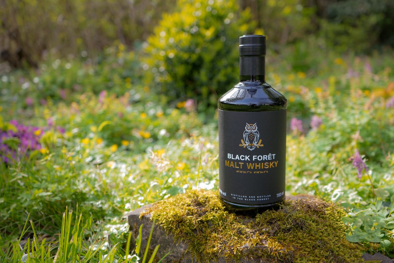 Eine Flasche Black Forêt Malt Whisky steht auf Moos in einem blühenden Garten.