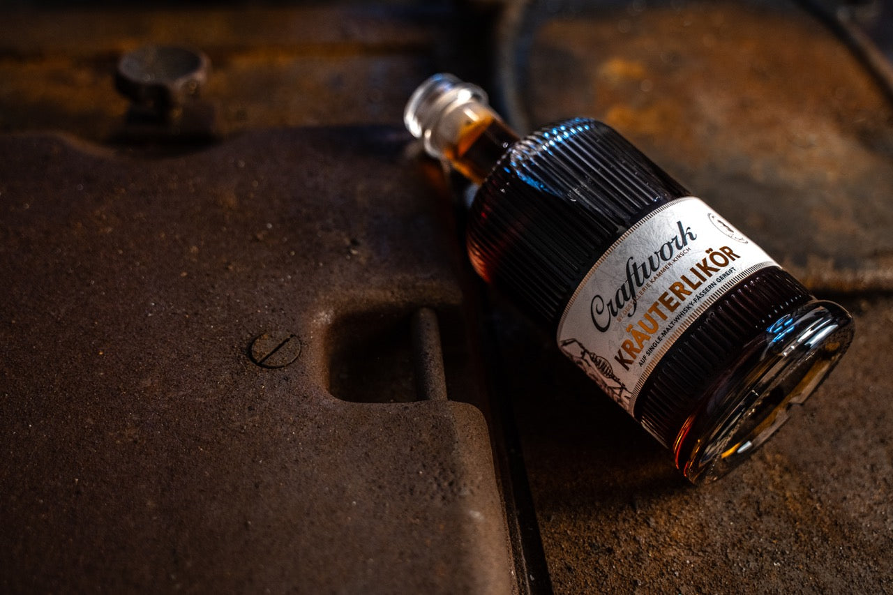 Eine Flasche Kräuterlikör mit Etikett "Craftwork" liegt auf einer rustikalen, metallischen Oberfläche.