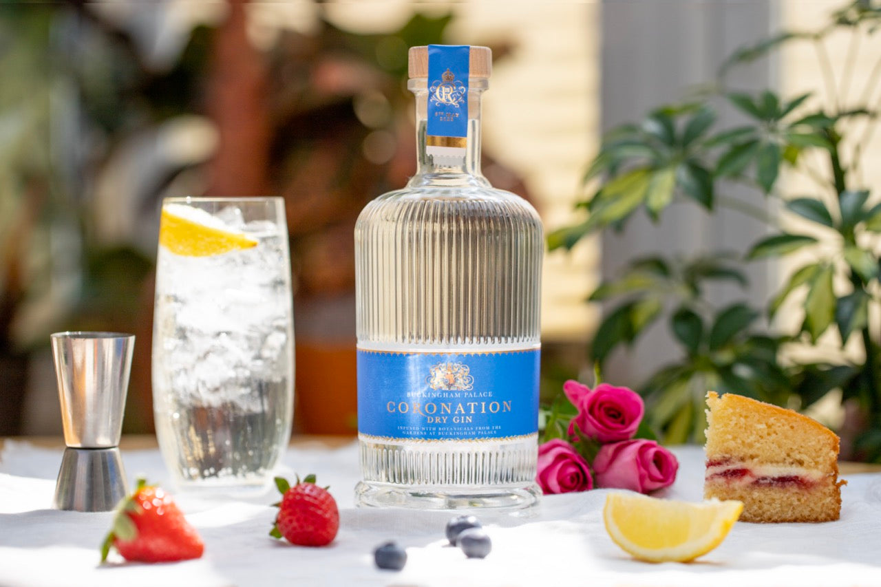 Eine Flasche Coronation Dry Gin mit einem Cocktailglas, Obst, Kuchenstück und rosa Rosen auf einem Tisch.