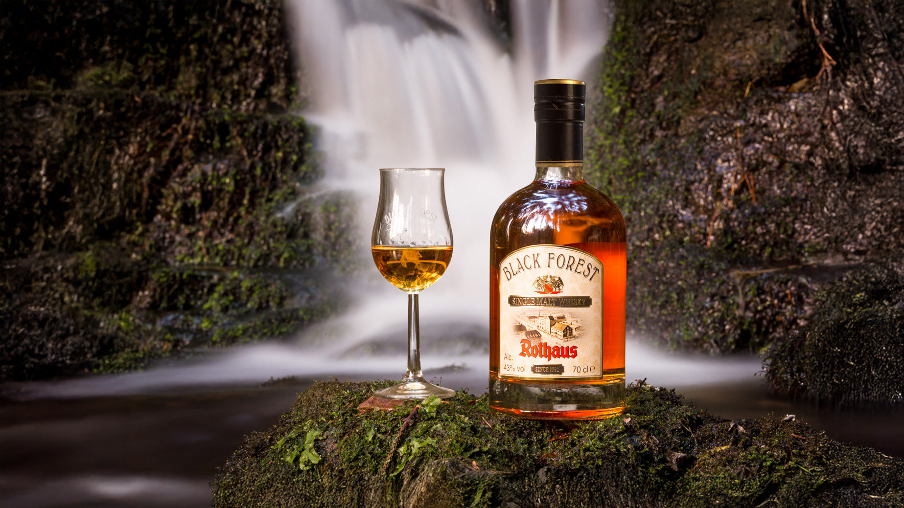 Eine Flasche Black Forest Whisky und ein gefülltes Glas stehen auf Moos vor einem Wasserfall.