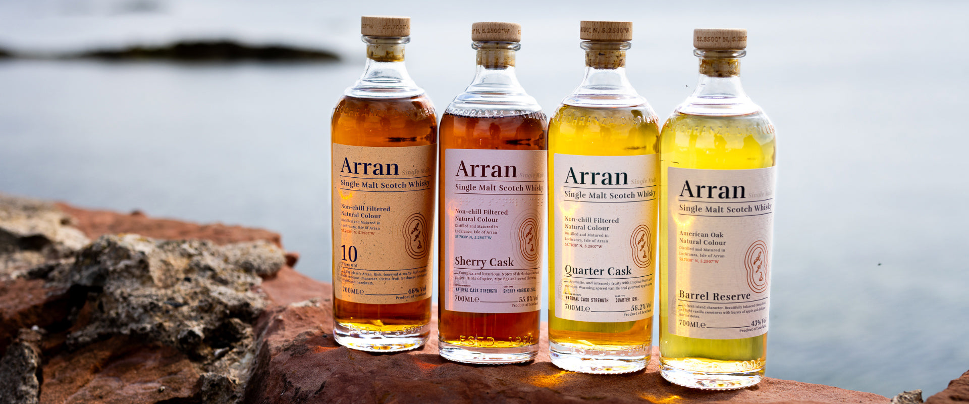 Vier Flaschen Arran Single Malt Scotch Whisky auf einem Felsen mit unscharfem Wasser im Hintergrund.
