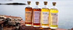 Vier Flaschen Arran Single Malt Scotch Whisky auf einem Felsen mit unscharfem Wasser im Hintergrund.