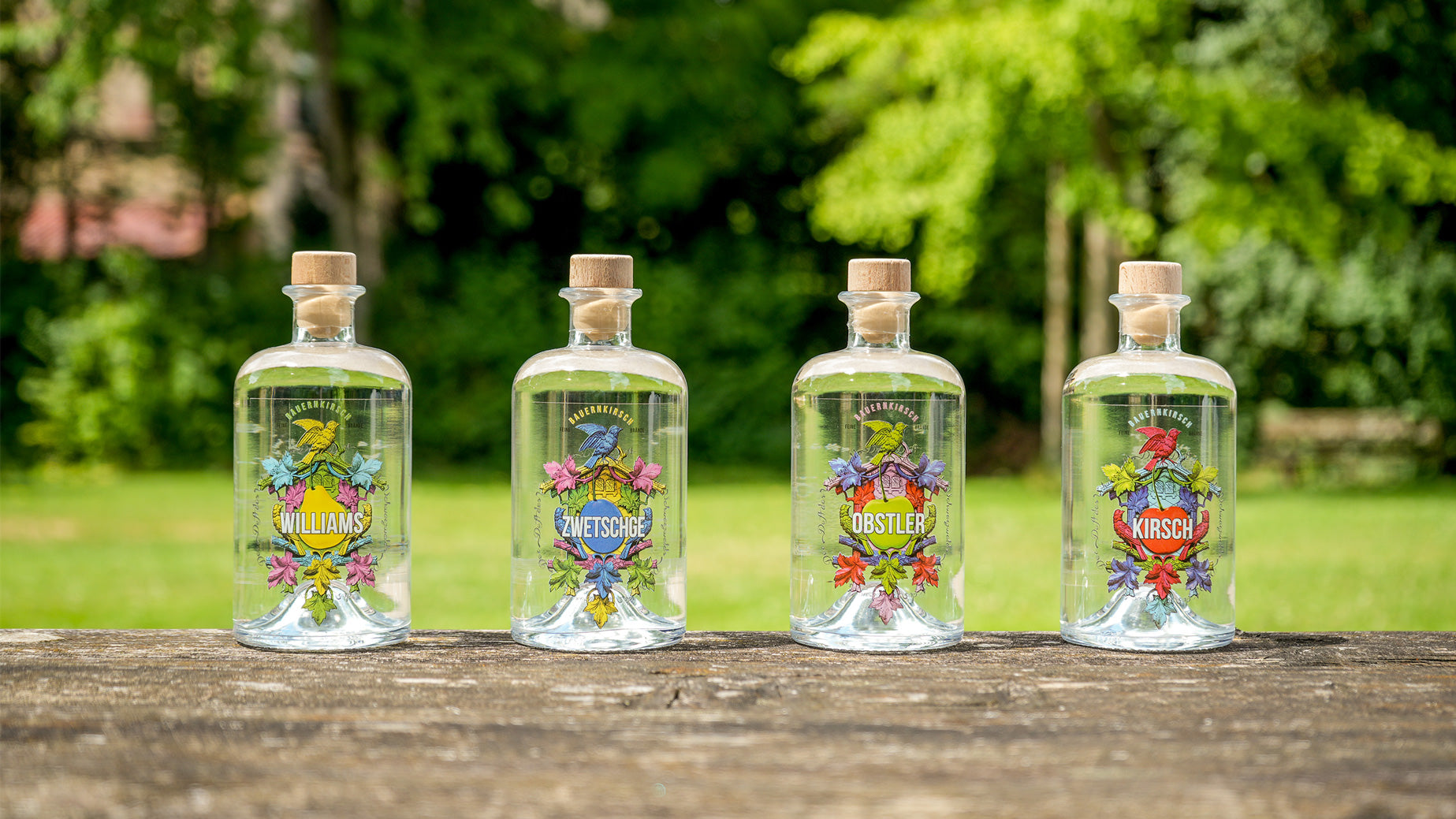 Vier Flaschen Obstbrand mit Etiketten "Williams", "Zwetschge", "Obstler" und "Kirsch" auf einem Holztisch im Freien.