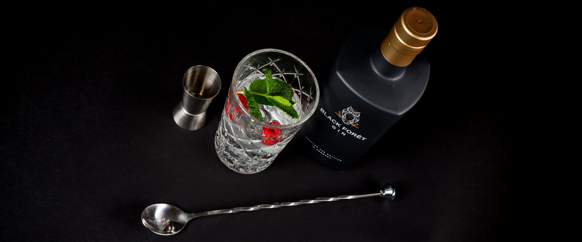 Flasche Gin, Glas mit Minze und Himbeeren, Messbecher und Barlöffel auf schwarzem Hintergrund.
