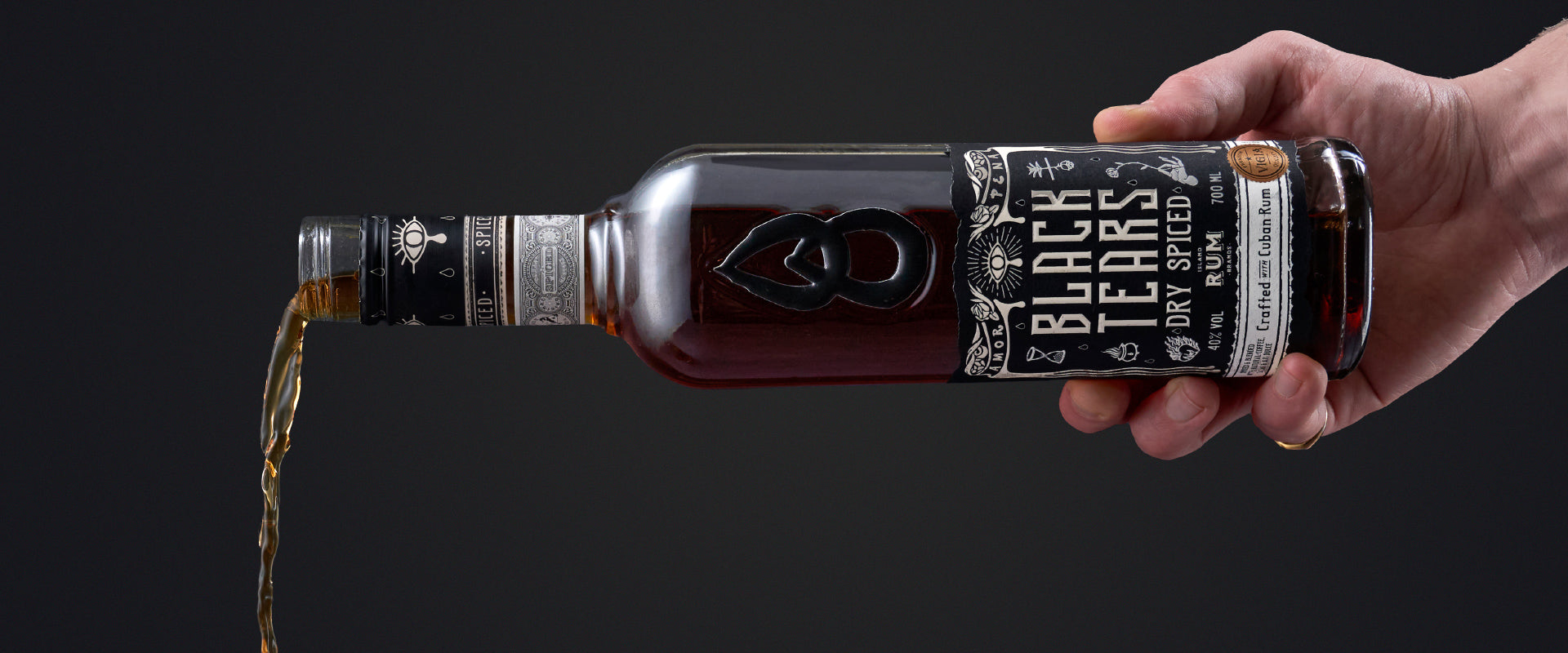 Eine Hand gießt flüssigen Inhalt aus einer Flasche "Black Tears Dry Spiced Rum" vor dunklem Hintergrund.