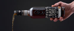 Eine Hand gießt flüssigen Inhalt aus einer Flasche "Black Tears Dry Spiced Rum" vor dunklem Hintergrund.