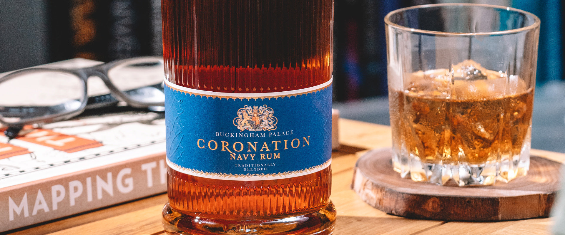 Eine Flasche Buckingham Palace Coronation Navy Rum neben einem Glas mit Rum und einem Buch mit Brille.