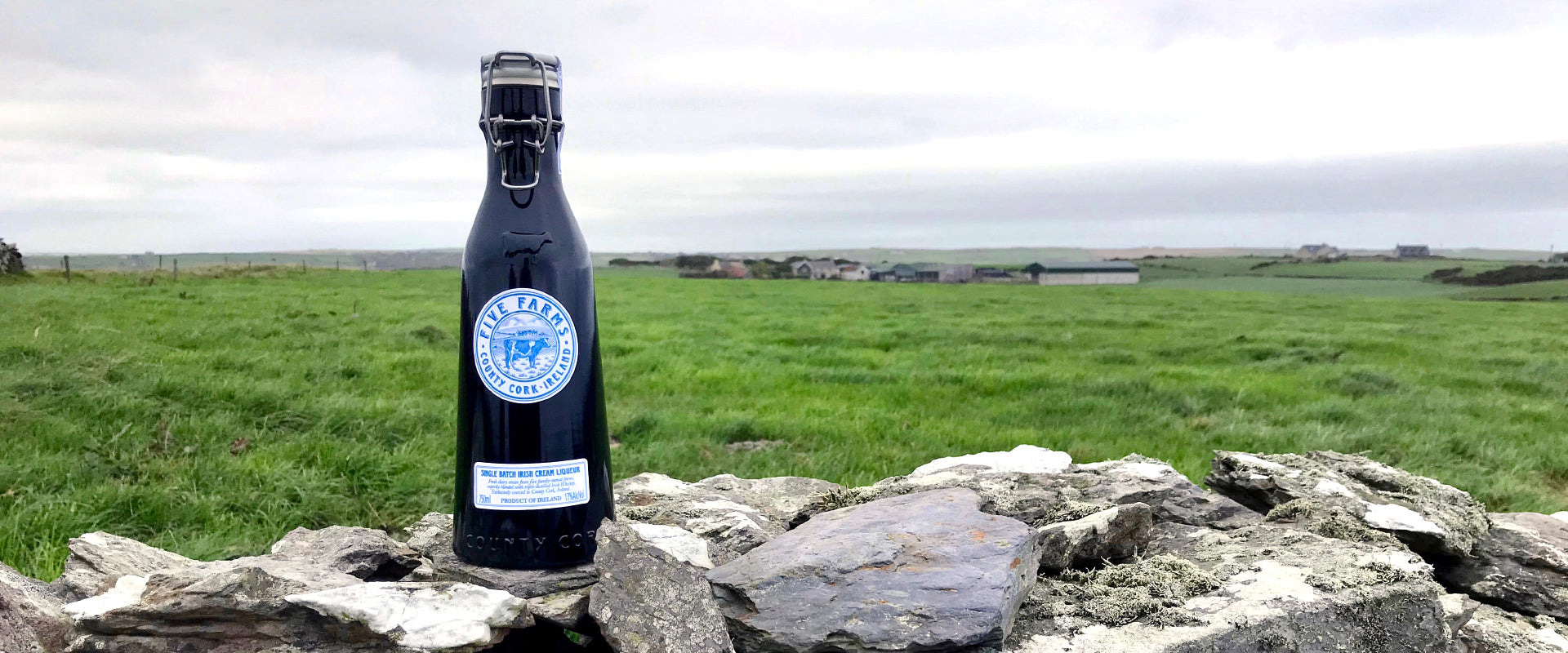 Eine schwarze Flasche mit "Five Farms"-Logo steht auf einer Steinmauer vor einer grünen Wiesenlandschaft.