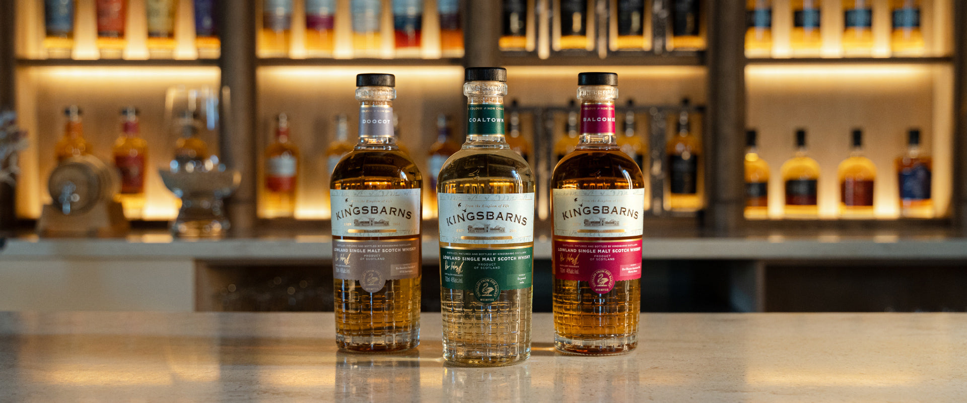 Drei Flaschen Kingsbarns Whisky auf einer Theke, im Hintergrund beleuchtetes Regal mit weiteren Flaschen.