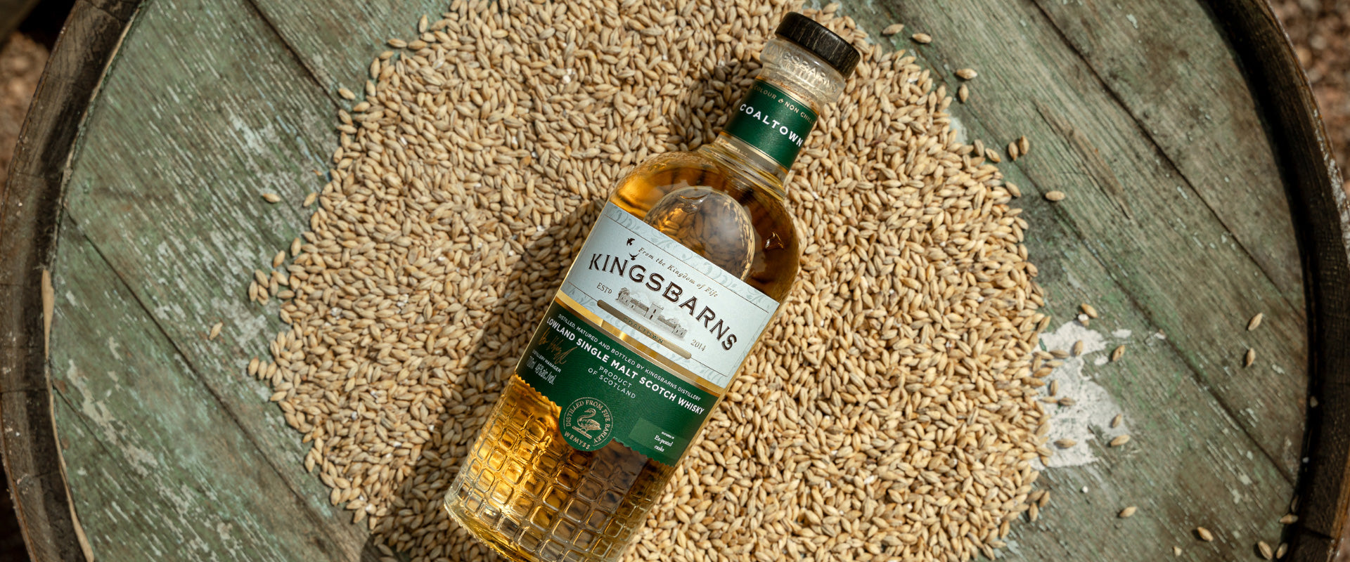 Flasche Kingsbarns Whisky liegt auf einem alten Holzfass, umgeben von Getreidekörnern.
