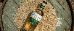 Flasche Kingsbarns Whisky liegt auf einem alten Holzfass, umgeben von Getreidekörnern.