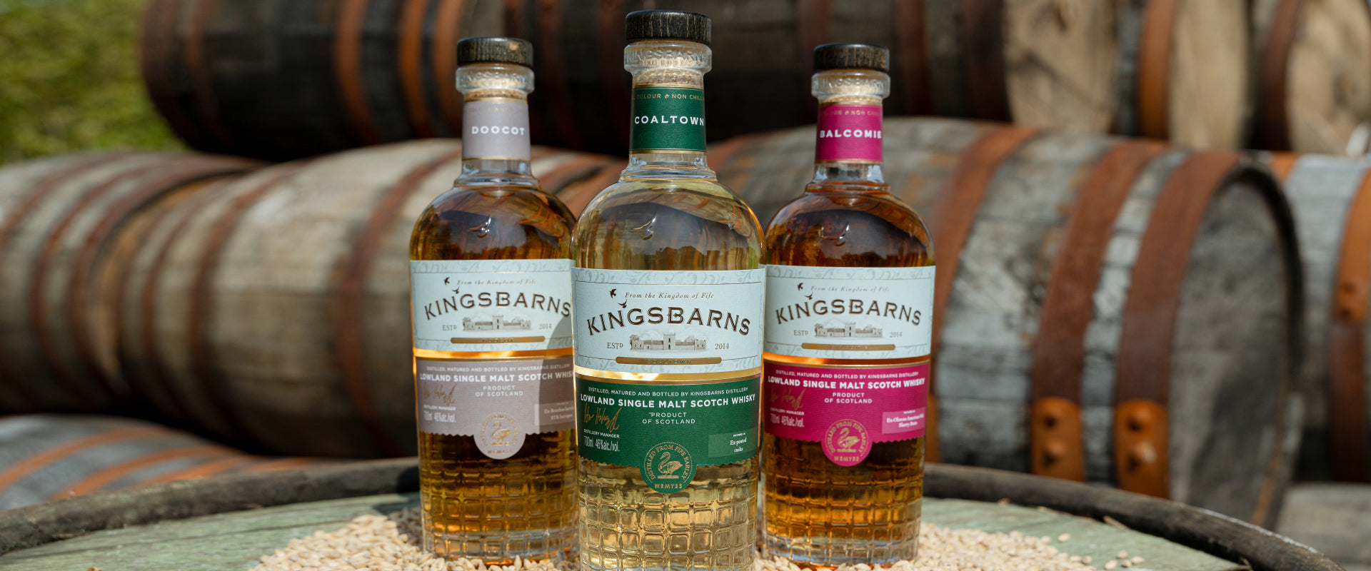 Drei Whiskyflaschen der Marke Kingsbarns stehen vor alten Holzfässern auf einem Tisch.