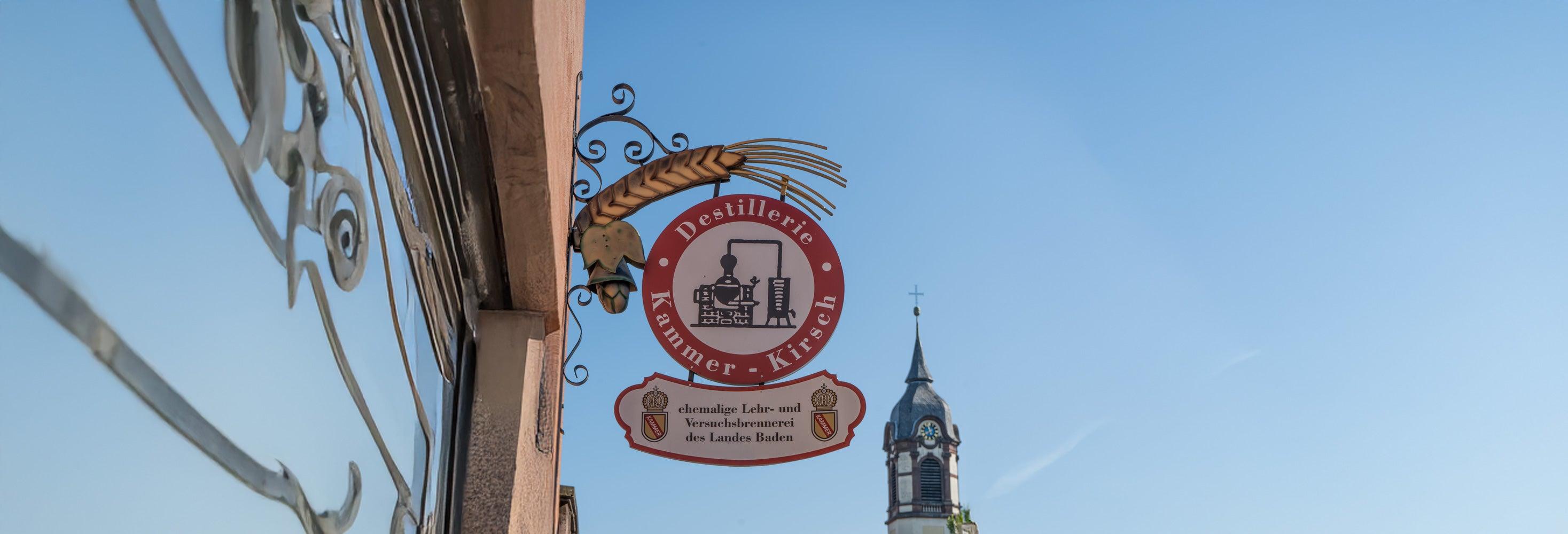Ein Schild einer Destillerie hängt an einer Hauswand, im Hintergrund ein Kirchturm und blauer Himmel.