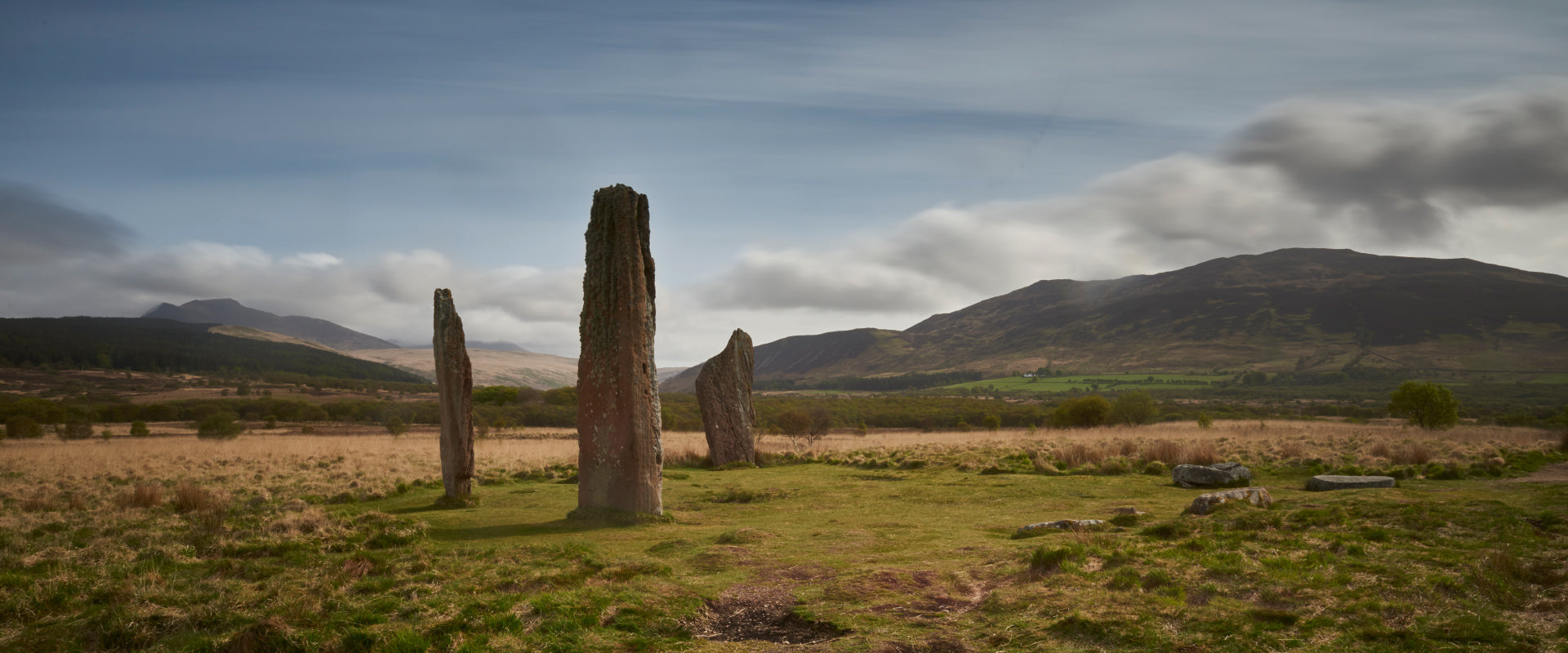 Graslandschaft mit stehenden Steinen und Bergen im Hintergrund unter teilweise bewölktem Himmel.