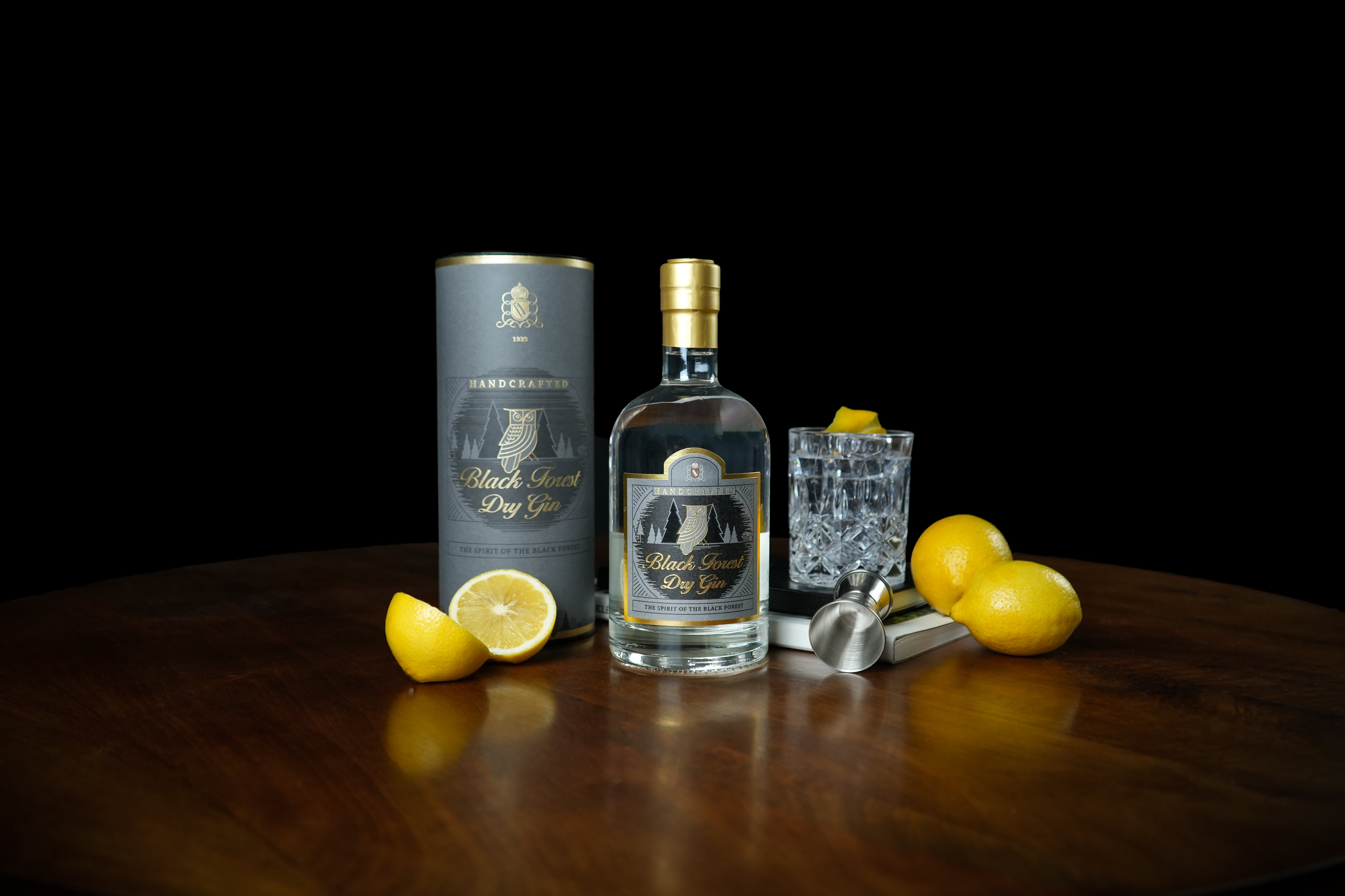 Eine Flasche Black Forest Dry Gin neben einem Cocktailglas, Zitronen und einem Messbecher auf einem Holztisch.