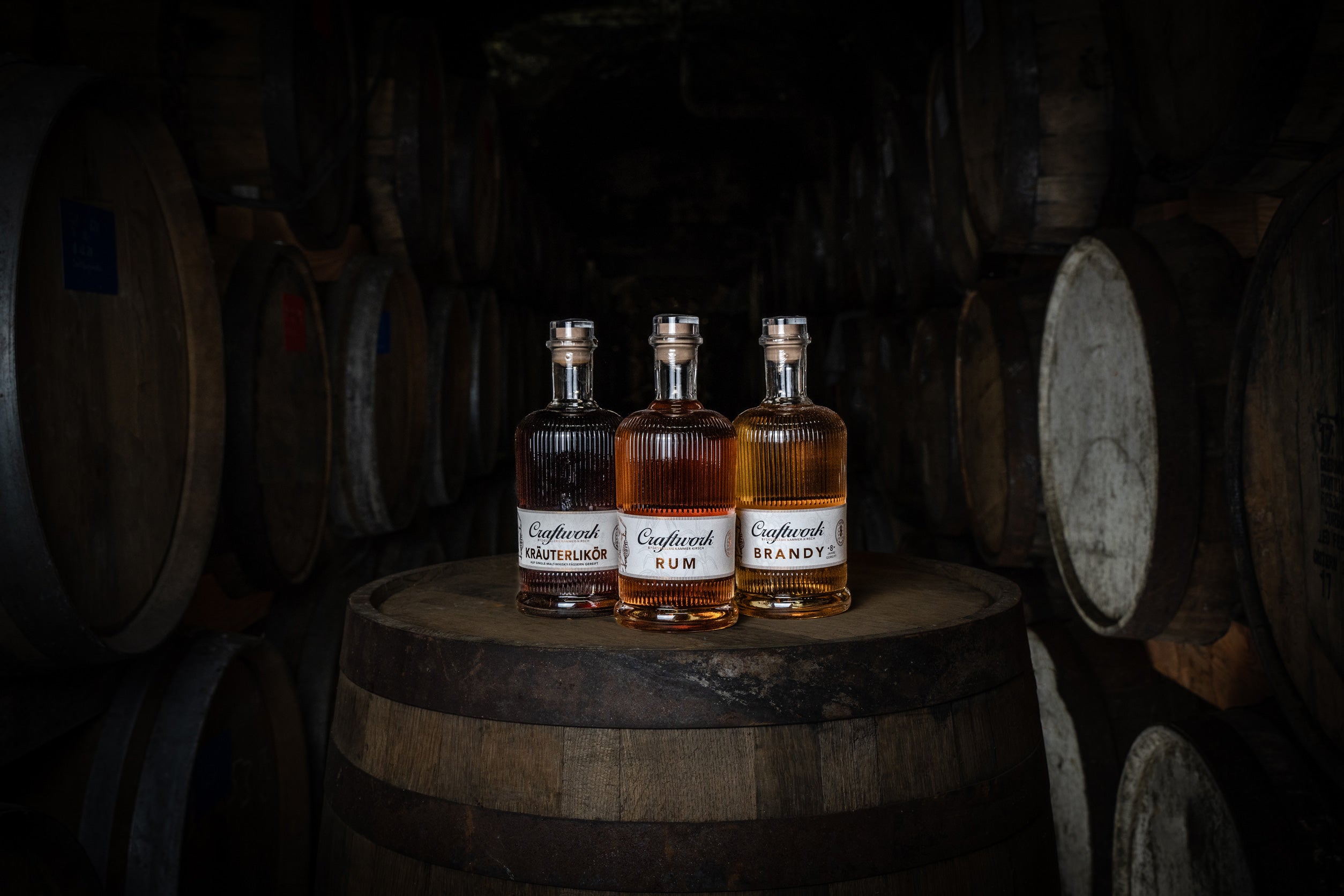 Drei Flaschen mit Alkohol stehen auf einem Holzfass in einem Keller mit Fässern im Hintergrund.