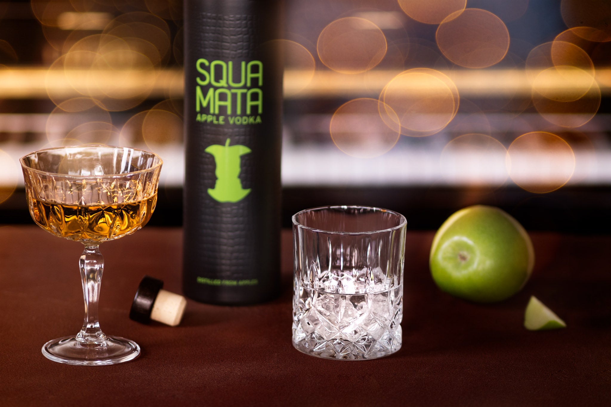 Zwei Gläser mit Getränken und eine Flasche Vodka, daneben ein grüner Apfel und ein Apfelstück auf einem Tisch.