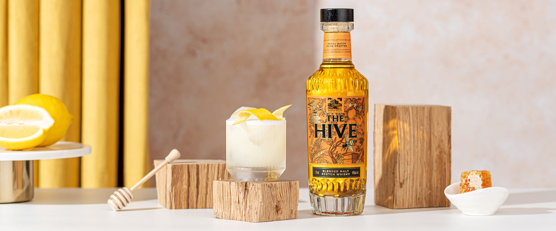 Flasche "The Hive" Whisky neben einem Cocktail mit Zitronengarnitur und einer Honigwabe auf weißem Tisch.
