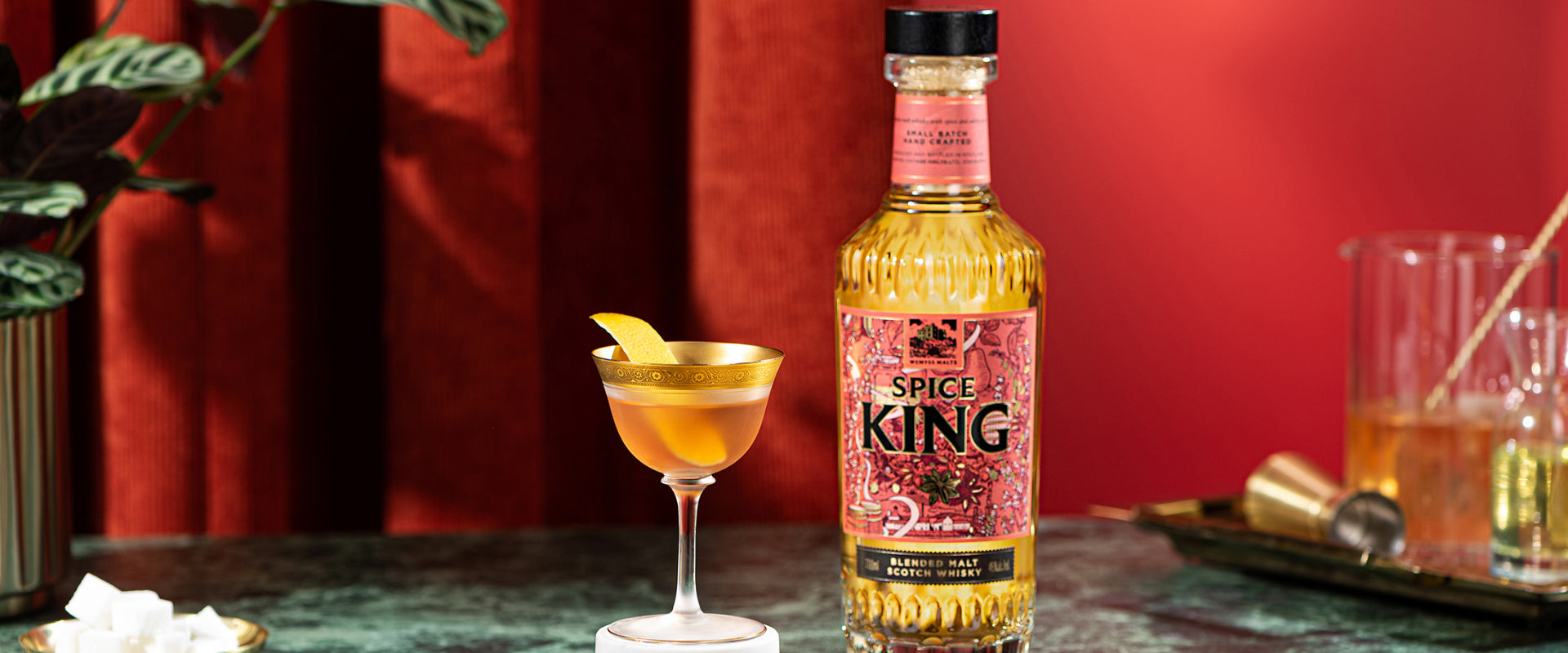 Eine Flasche "Spice King" Whisky neben einem Cocktailglas mit Orangenschale, vor rotem Hintergrund.