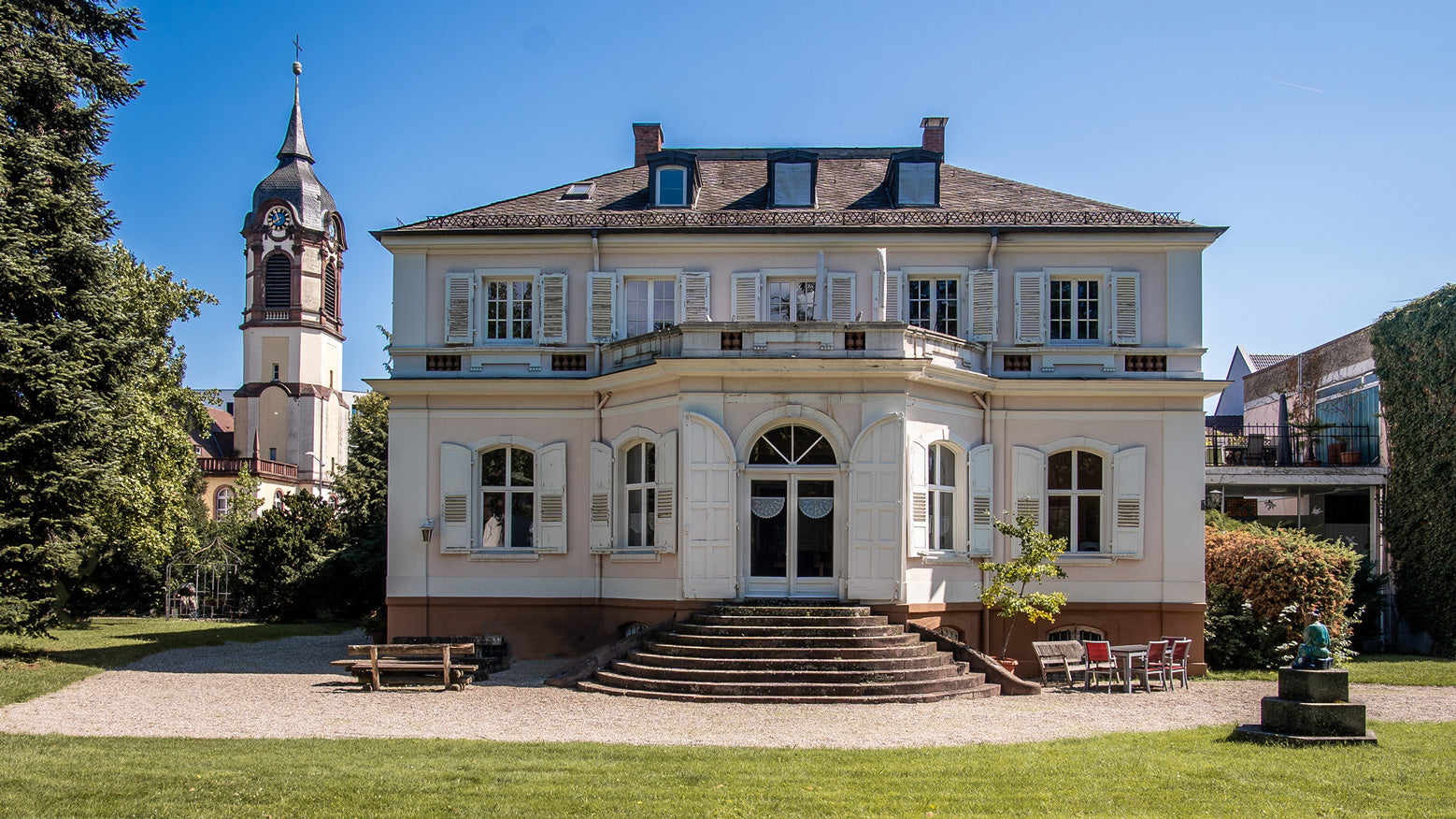 Historische Villa mit Garten und Turm einer Kirche im Hintergrund an einem sonnigen Tag.