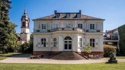Historische Villa mit Garten und Turm einer Kirche im Hintergrund an einem sonnigen Tag.