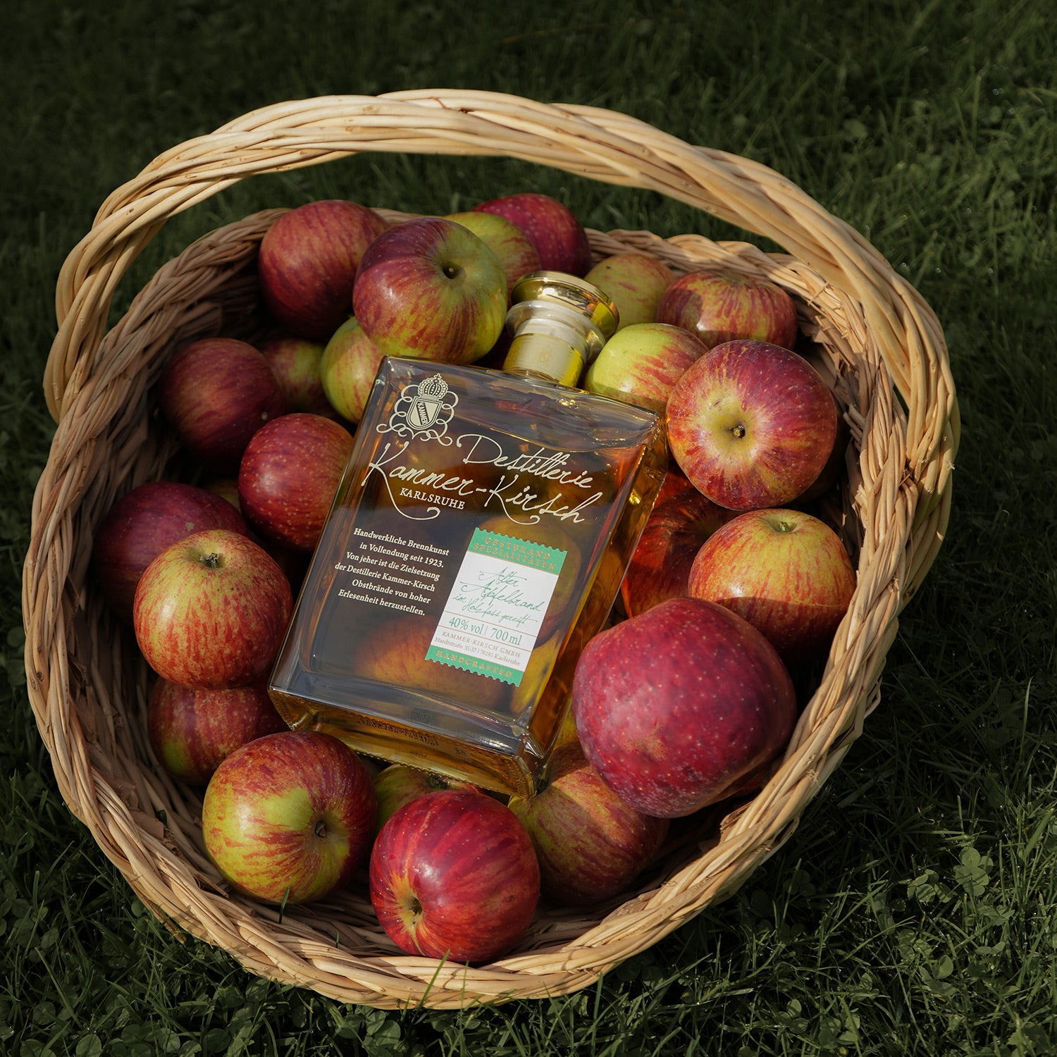 Korb mit roten Äpfeln und einer Flasche alkoholisches Getränk auf einer Wiese.