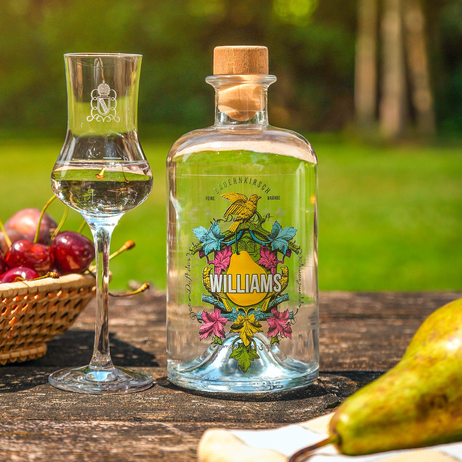 Eine Flasche Williams-Birnenbrand steht auf einem Tisch neben einem Glas und einem Korb mit Kirschen im Freien.