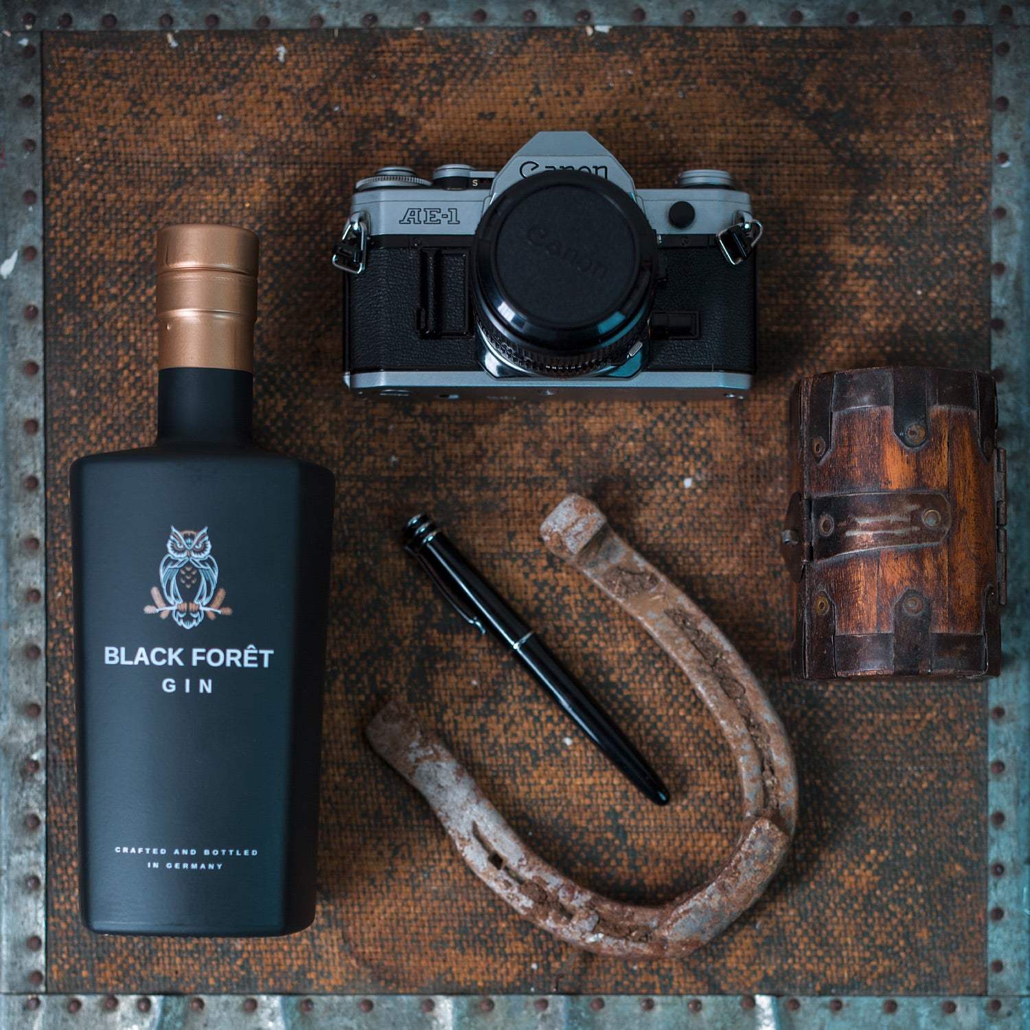 Eine Flasche Gin, eine alte Kamera, ein Hufeisen, ein schwarzer Stift und eine kleine Holztruhe auf rostigem Untergrund.