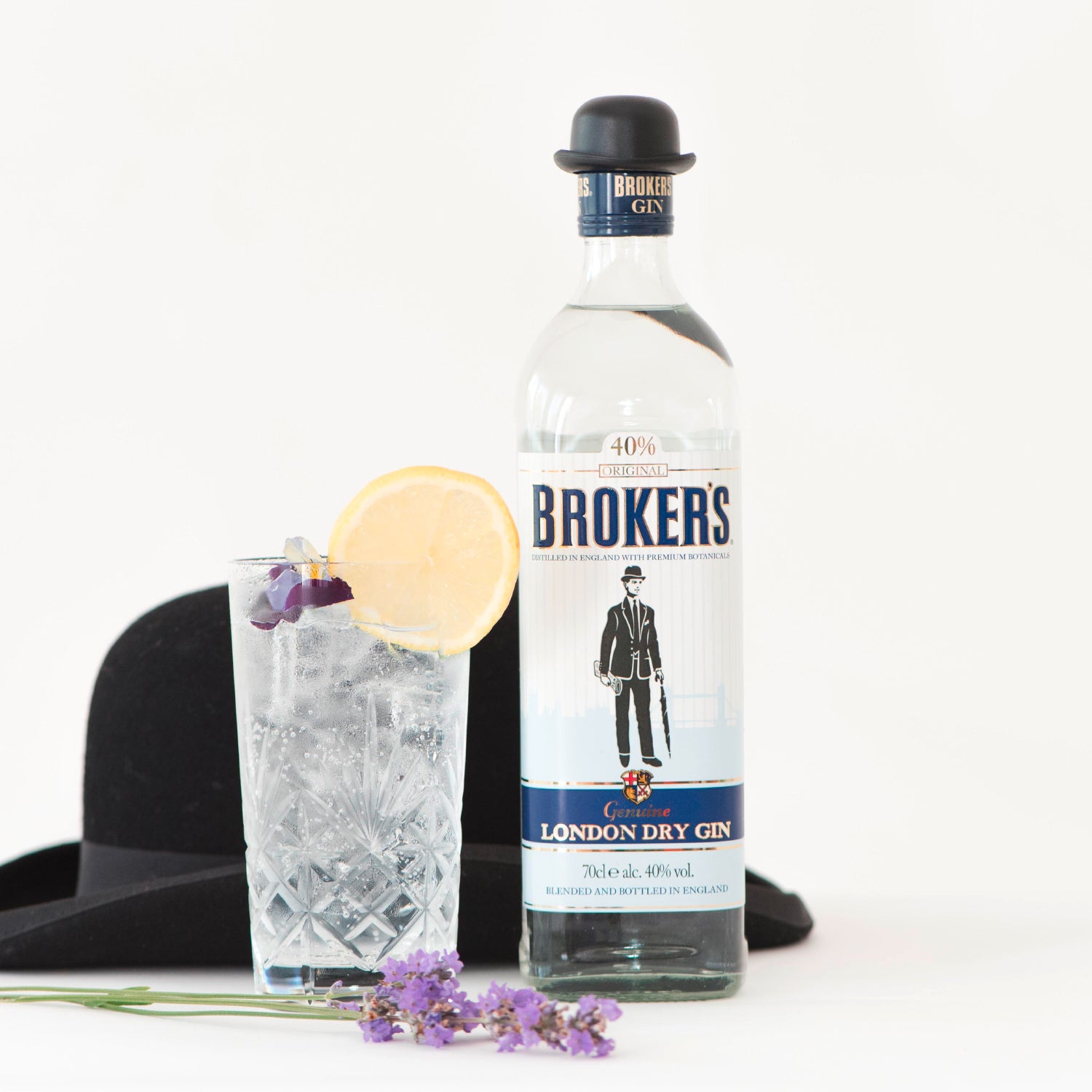 Eine Broker's Gin-Flasche neben einem gefüllten Glas mit Zitronenscheibe, einem schwarzen Hut und Lavendelblüten.