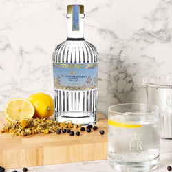 Buckingham Palace Dry Gin-Flasche mit Zitrone, Kamille und Wacholderbeeren auf Holzbrett, daneben ein Glas mit Gin Tonic.