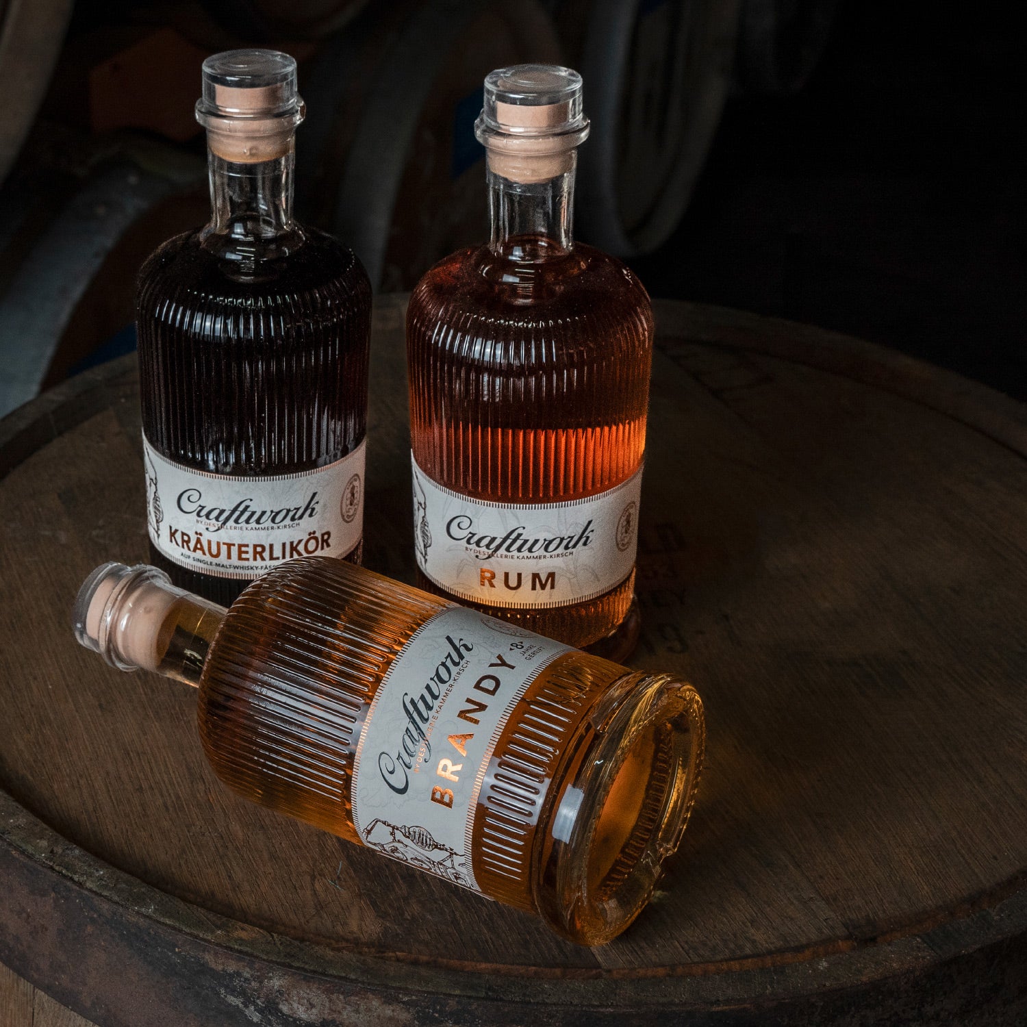 Drei Flaschen Craftwork Spirituosen auf einem Holzfass, darunter Kräuterlikör, Rum und Brandy.