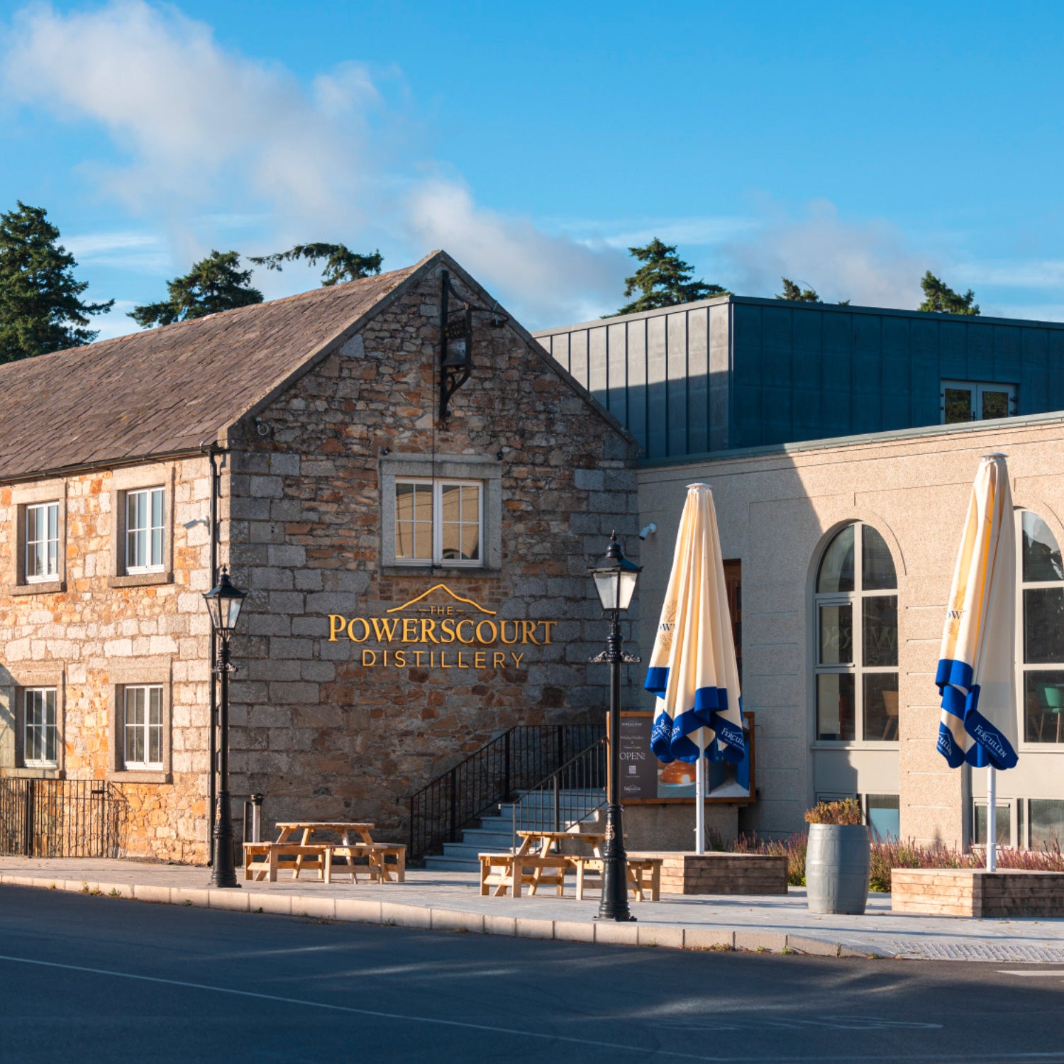 Außenansicht der Powerscourt Distillery mit Steinmauern, großen Fenstern und Sonnenschirmen im Vordergrund.