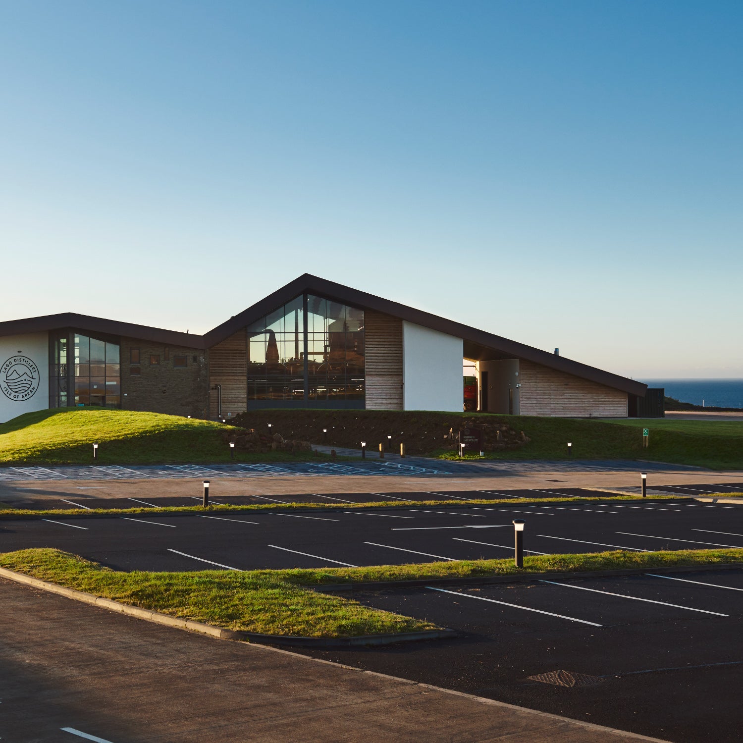 Ein modernes Gebäude mit großen Glasfronten und einem geneigten Dach, umgeben von einem leeren Parkplatz und grünem Rasen.