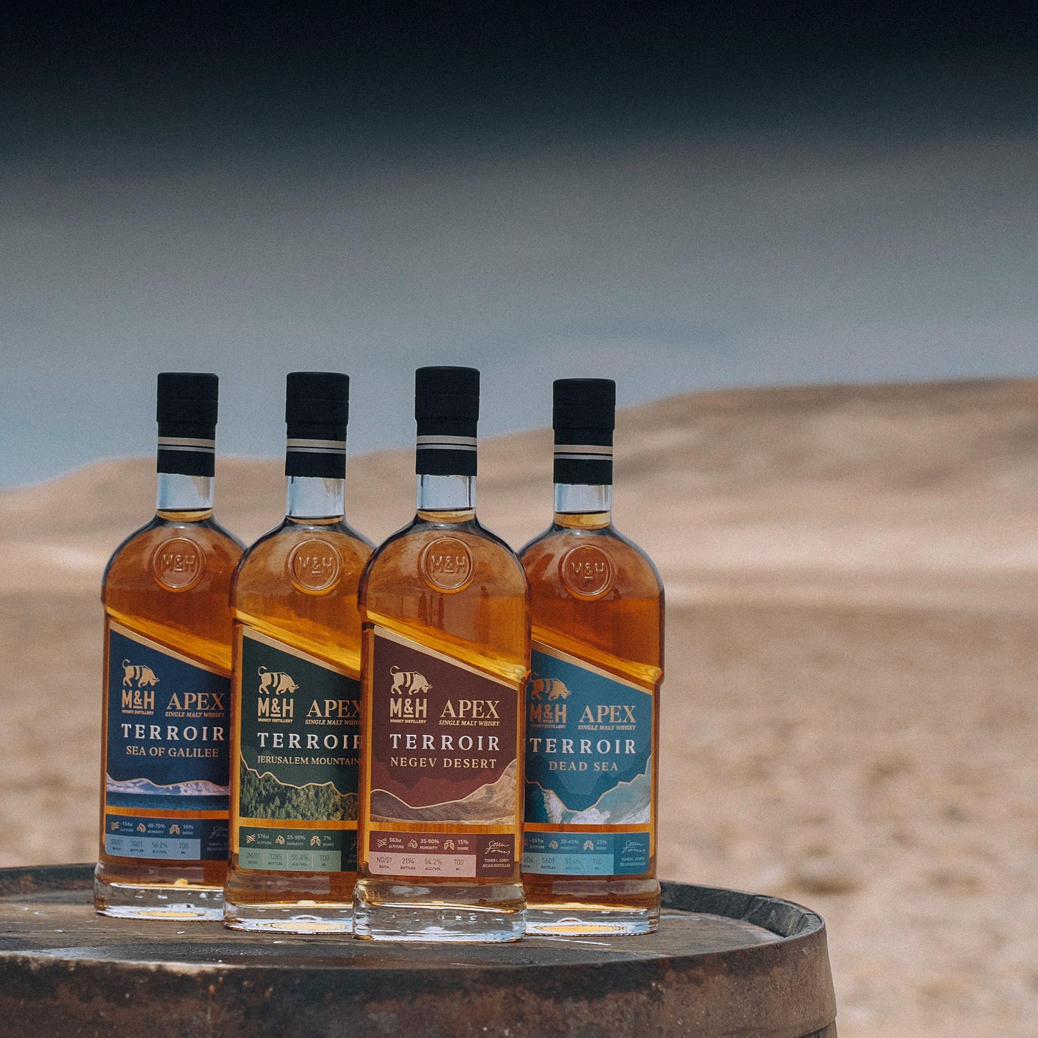 Vier Whiskyflaschen der Marke M&H mit unterschiedlichen Etiketten stehen auf einem Fass in einer Wüstenlandschaft.