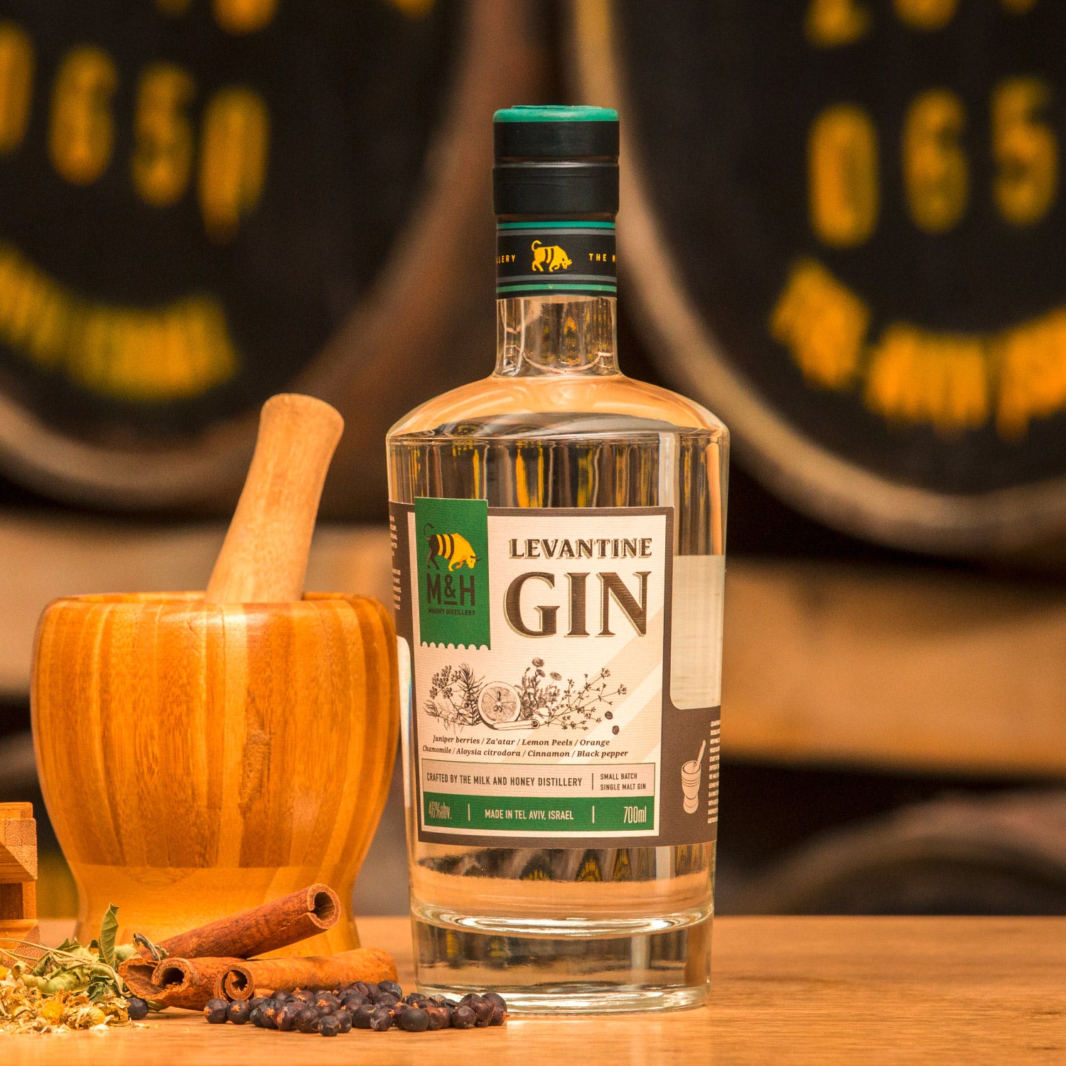 Eine Flasche Levantine Gin steht neben einem Holzstößel und verschiedenen Gewürzen auf einem Tisch vor einem Barriquefass.